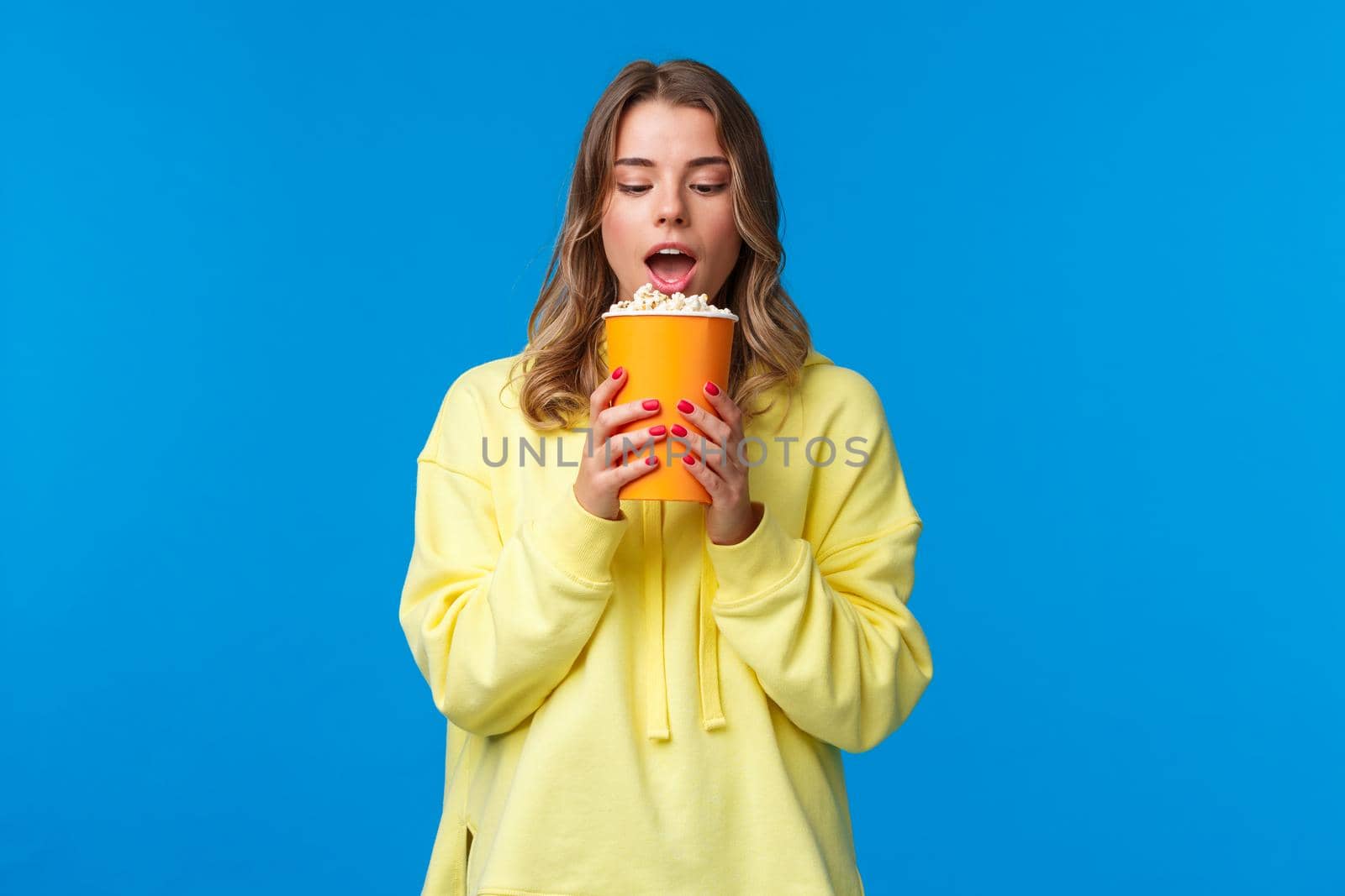 Leisure, fun and youth concept. Girl likes eating popcorn in cinema, have bite and staring at can with desire, watching movie, standing blue background in yellow hoodie. Copy space