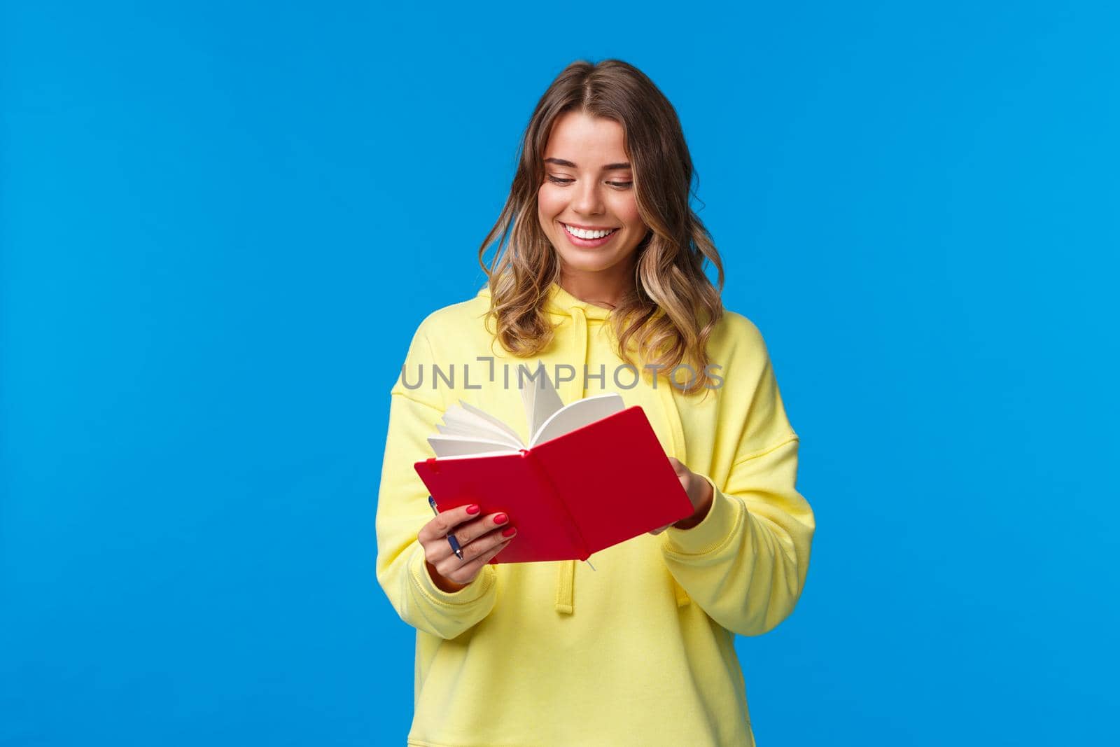 Girl bought new planner, writing down notes or lecture, smiling joyfully, holding red notebook, decide start secret diary, standing in yellow hoodie over blue background.