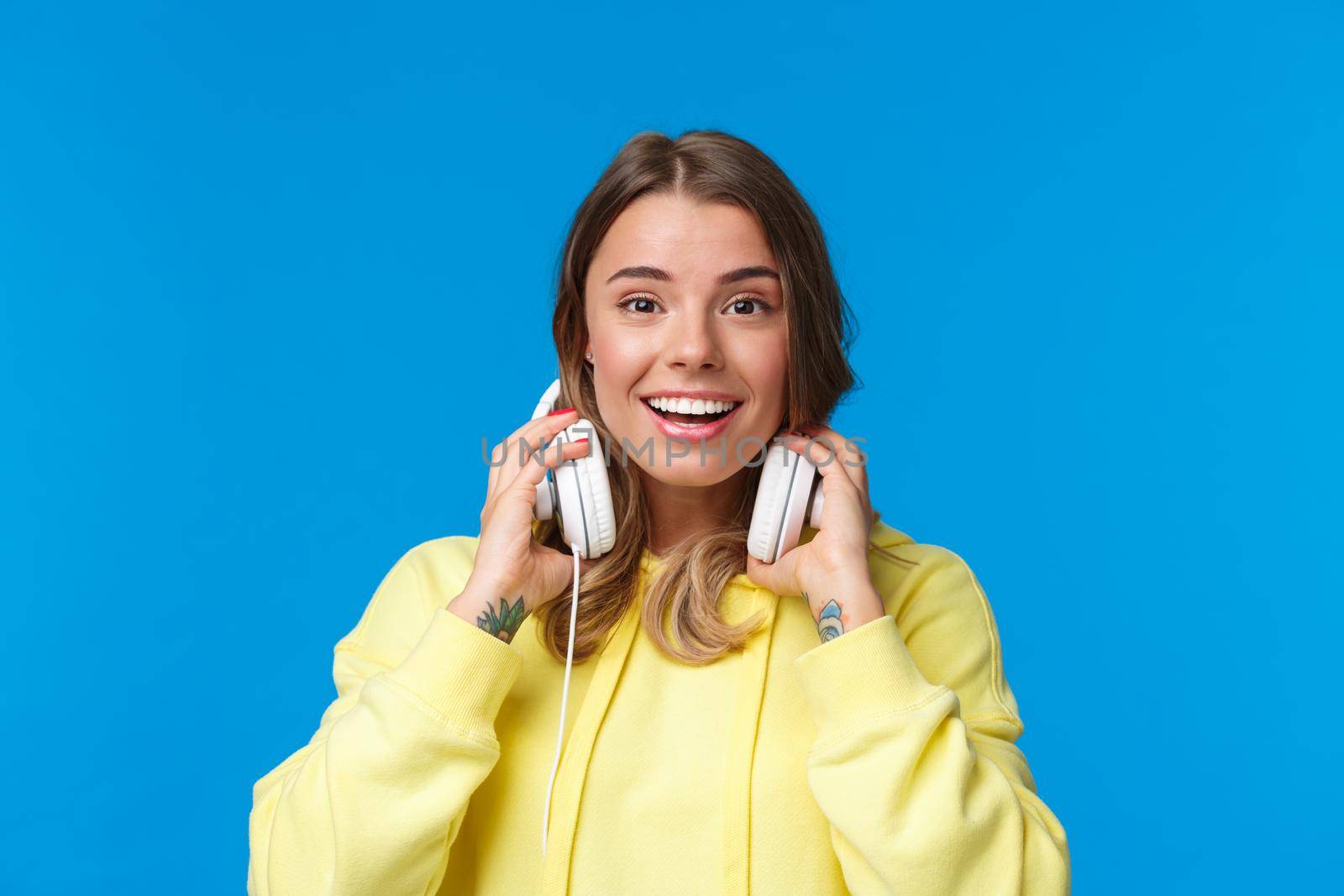 Close-up portrait of excited happy smiling blond girl in yellow hoodie, take-off headphones and grinning as meet friend on street stop by for conversation, standing joyful blue background.