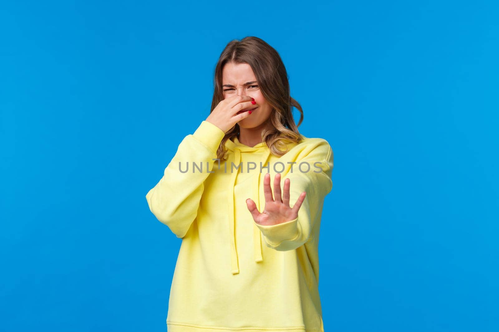 Oh gosh take this away from my face. Bothered and disturbed young blond woman close her nose and grimacing from awful nasty smell, pulling hand in stop refusal gesture, blue background.