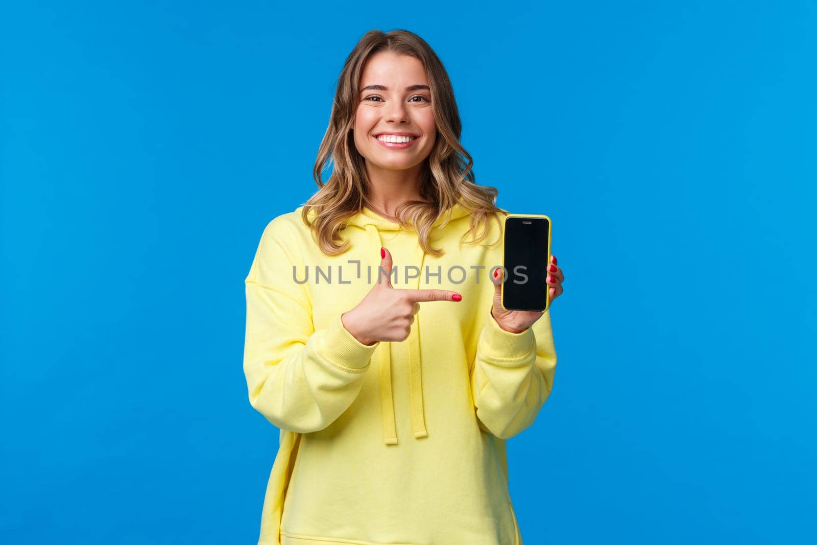 Proud cheerful young blond woman advertise new mobile game or application, look camera and smiling white teeth as pointing smartphone display, recommend download or try out by Benzoix