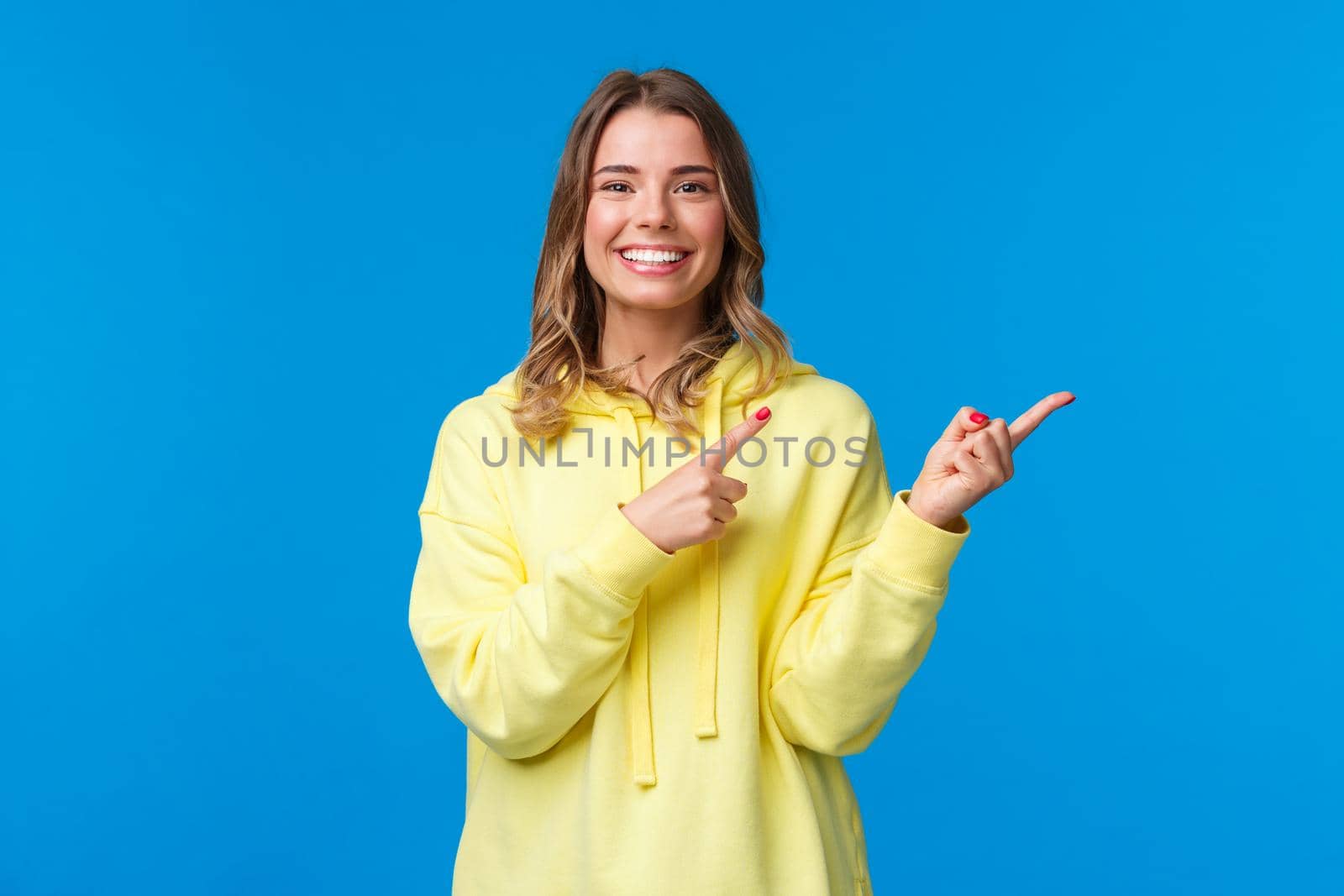 Good-looking friendly pleasant blond girl in yellow hoodie pointing fingers right and smiling white teeth at camera as recommend place, asking visit or click link, standing blue background.