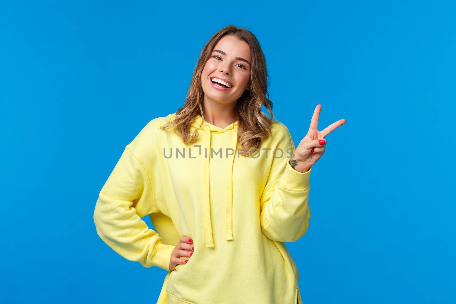 Staying bright sie. Carefree happy young silly blond girl in yellow hoodie sending positive vibes, show kawaii peace sign and smiling with white teeth, standing blue background.