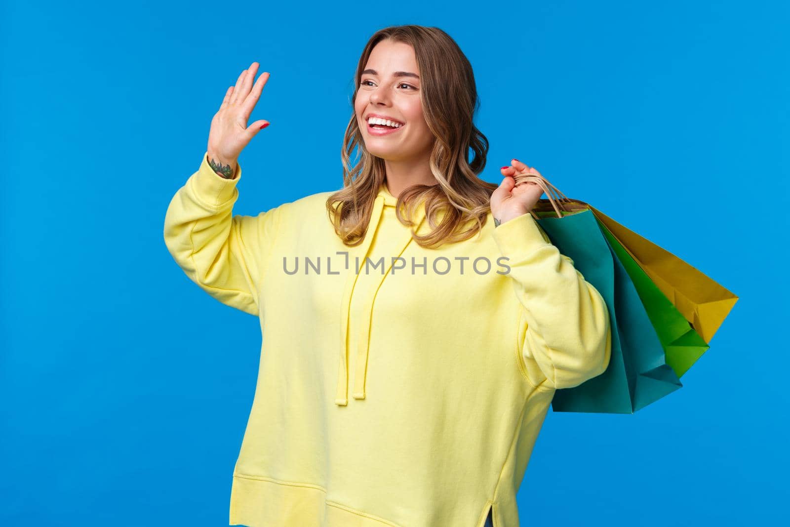 Cheerful happy and relaxed young blond girl holding shopping bags behind and waving friend, smiling friendly say hello as see mate in mall, buying things for vacation, blue background.