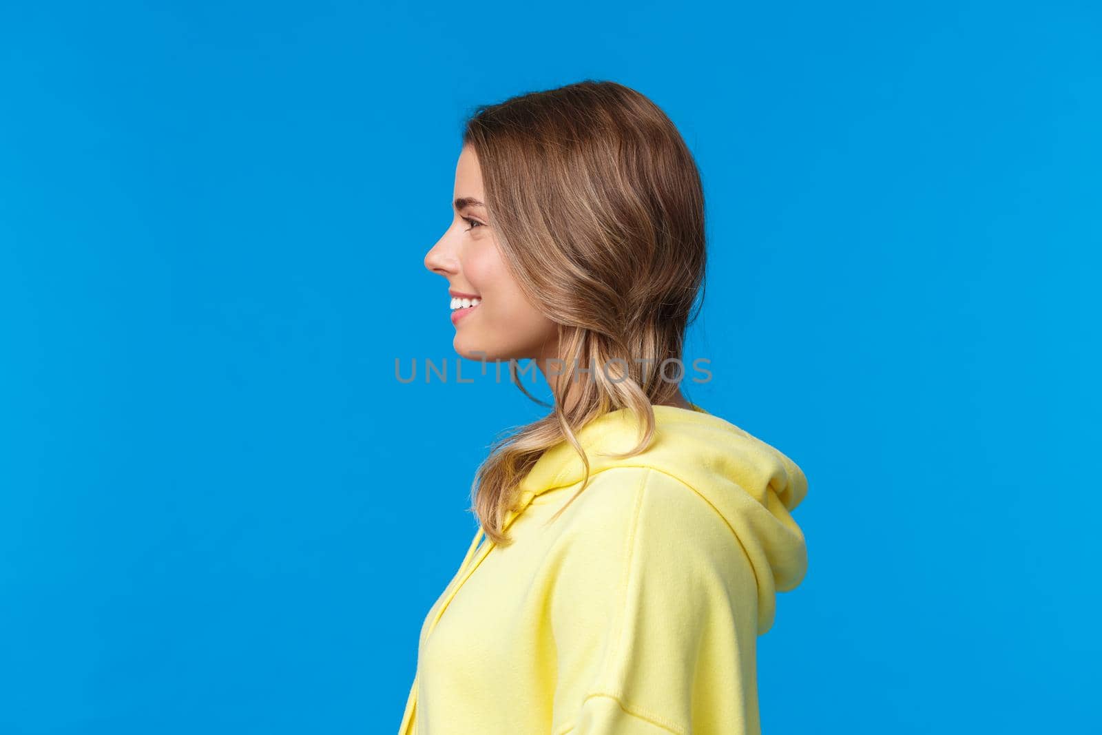 Close-up profile portrait of smiling cute young blond girl with short haircut, wearing yellow hoodie, facing left side of blank copy space, standing blue background upbeat. Copy space