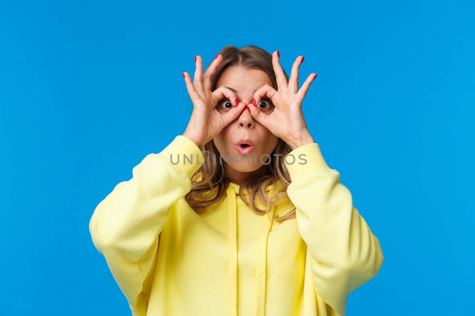 Impressed and intrigued, curious young caucasian girl in yellow hoodie looking at something awesome, found great product special discount, make glasses with fingers on eyes, blue background by Benzoix