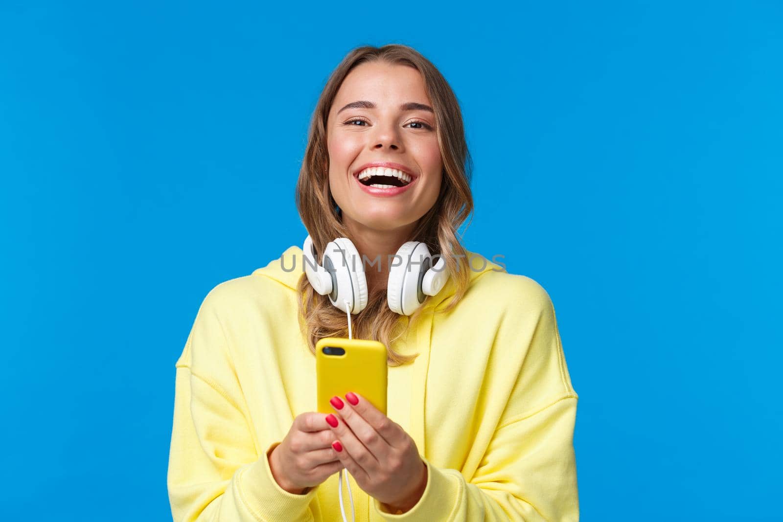 Close-up portrait joyful good-looking blond girl in yellow hoodie and white headphones, using mobile phone, writing in her blog, sharing news with social media, messaging and laughing camera.