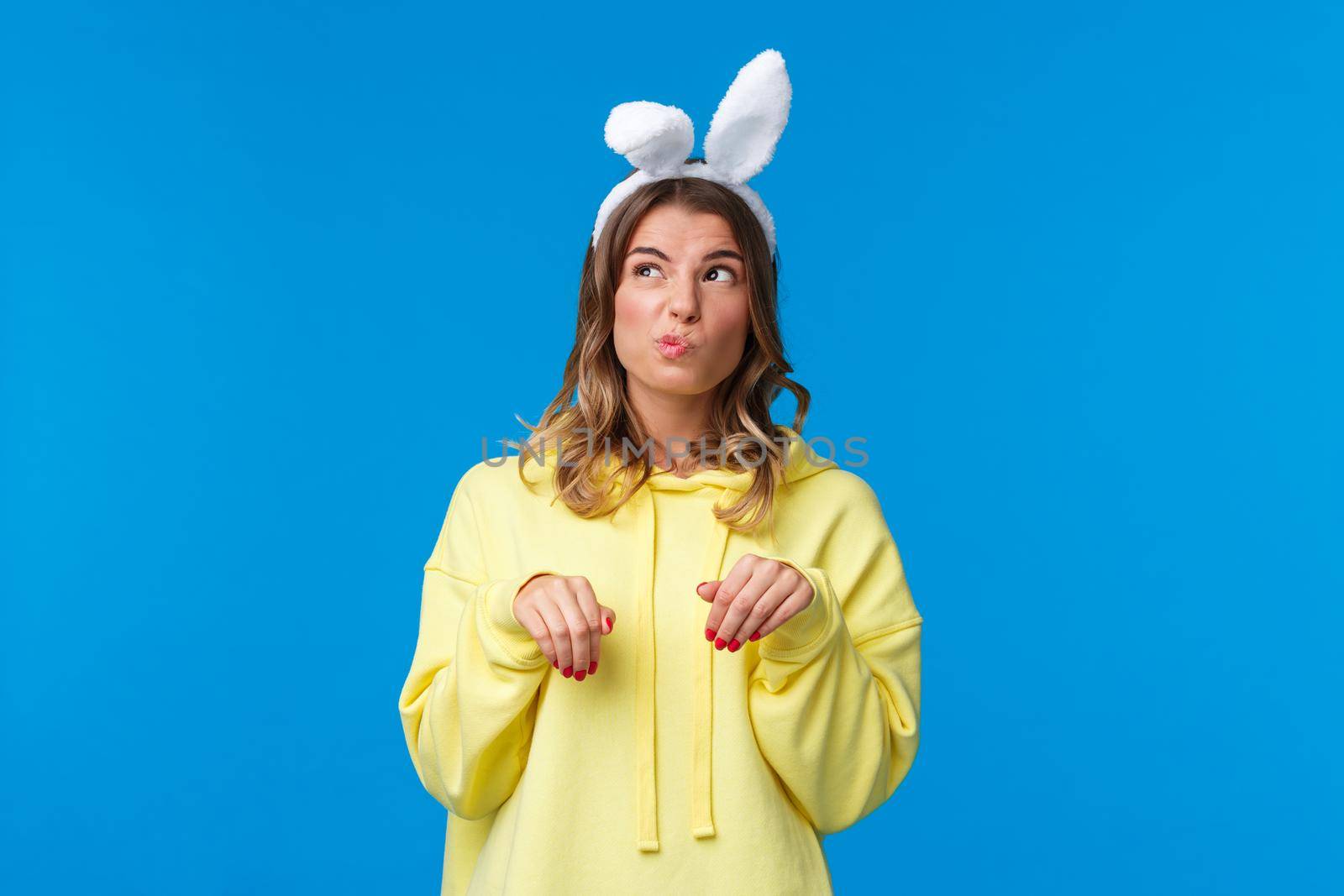 Thoughtful and cute funny blond girl acting like Easter rabbit with fake ears and paws gesture, pouting looking upper left corner mimicking bunny animal, stand blue background.