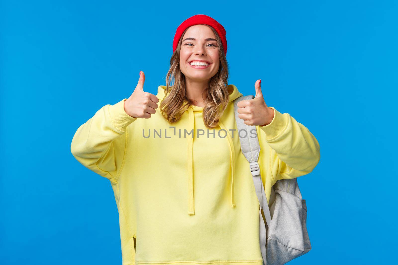 Sounds good, deal. Cheerful happy and smiling hipster girl in red beanie and yellow hoodie, approve idea, agree with friends picking where hang out after classes, hold backpack, blue background.
