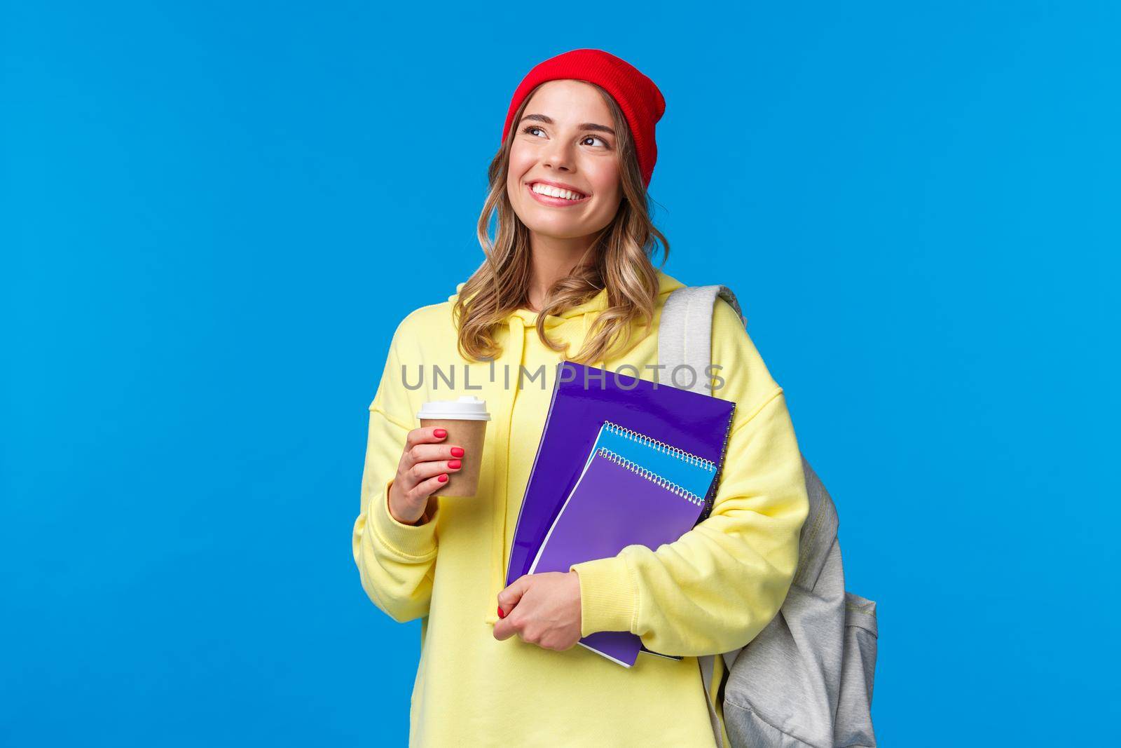 Dreamy cute college girl contemplating sunny spring day on her way to classes, holding take-away coffee and notebooks, smiling as looking up, carry backpack, standing blue background.