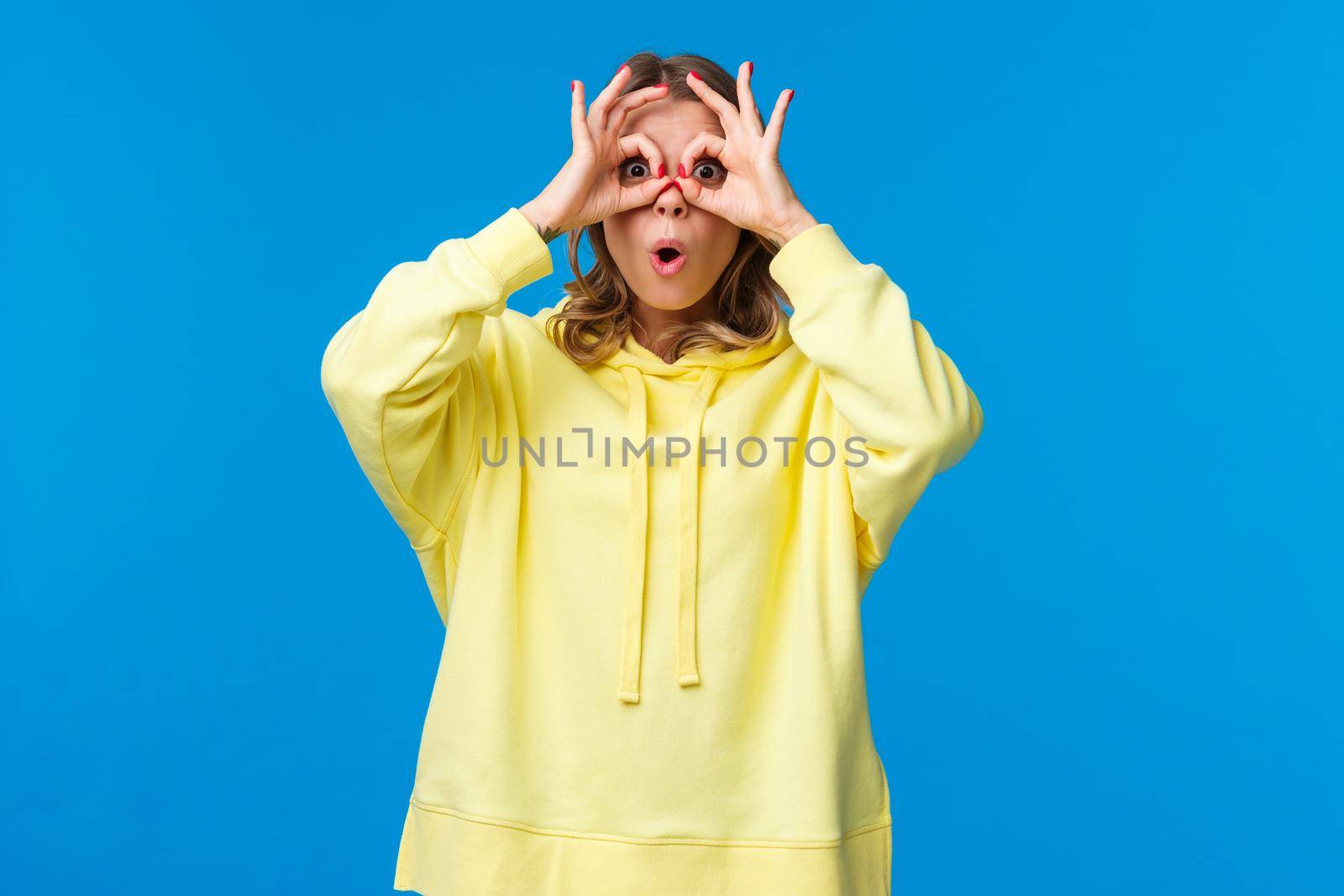 Wow interesting. Intrigued and surprised curious blond girl seeing something intriguing, open mouth astonished, look through hand-glasses and standing blue background.