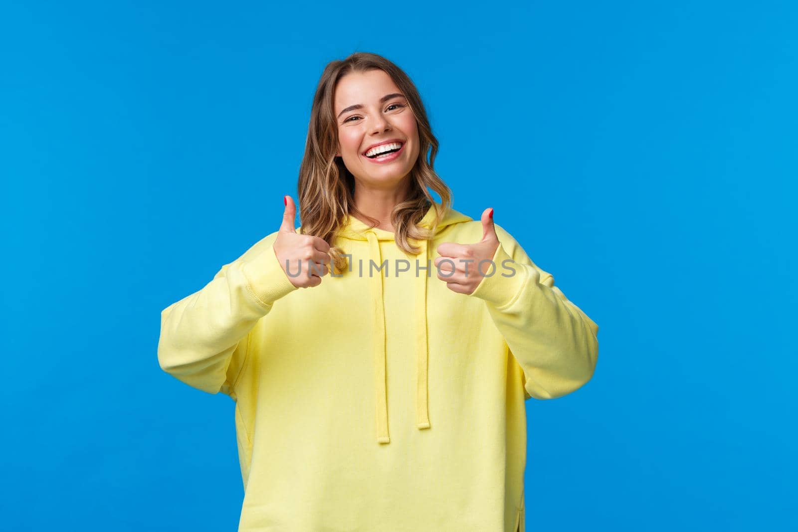 Sounds good Im in. Cheerful and optimistic good-looking young female blonde in yellow hoodie, show thumbs-up in approval, smiling nod acceptingly, like idea, blue background.