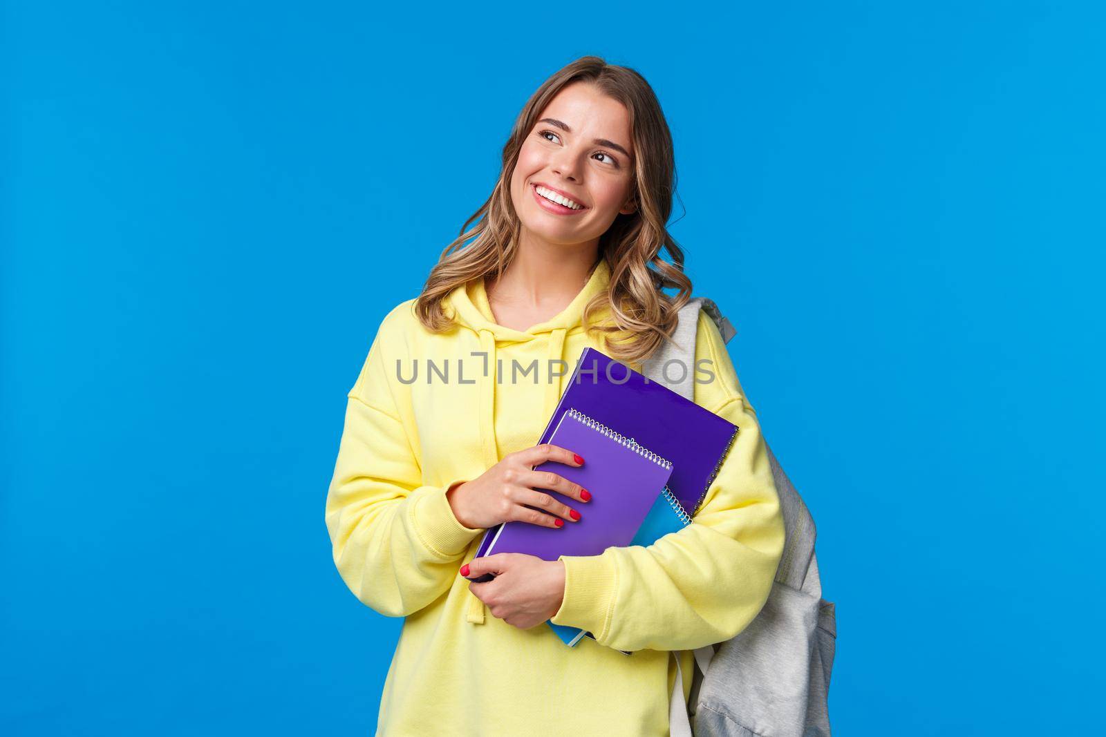 Dreamy happy good-looking pretty blond girl studying in college, holding backpack and notebooks, homework staff, look upper left corner thoughtful, standing blue background.