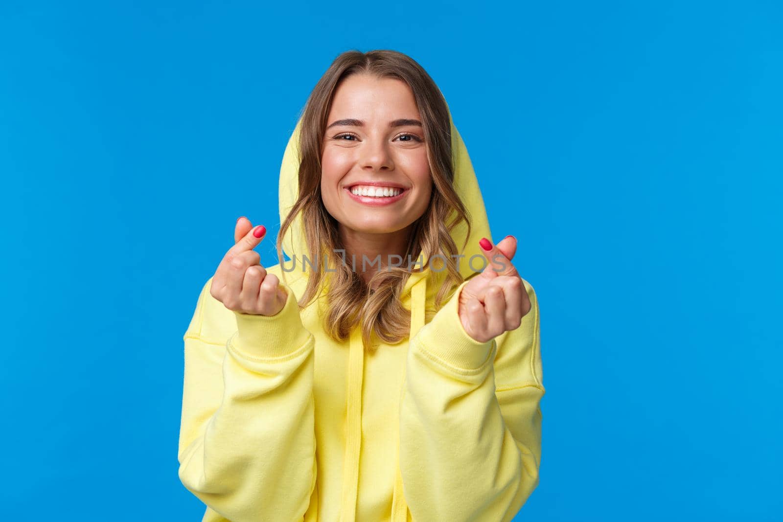 Close-up portrait of cute and silly blond caucasian girl in yellow hoodie, showing korean heart gesture and smiling, vibing, enjoying party, having fun over blue background. Copy space