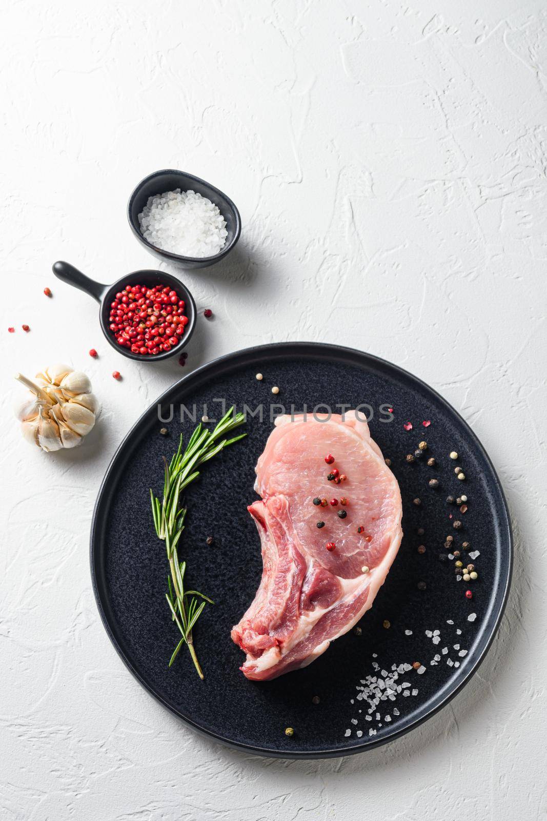 Raw Pork Loin chops in a black round plate on a white stone background with rosemary garlic peppercorns ingredients for grill top view vertical space for text.