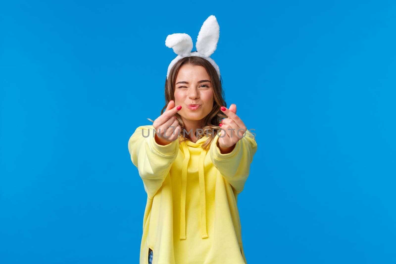 Holidays, traditions and celebration concept. Silly and cute happy blond girl having fun, showing korean heart gesture with fingers and send kisses, standing in rabbit ears, yellow hoodie.