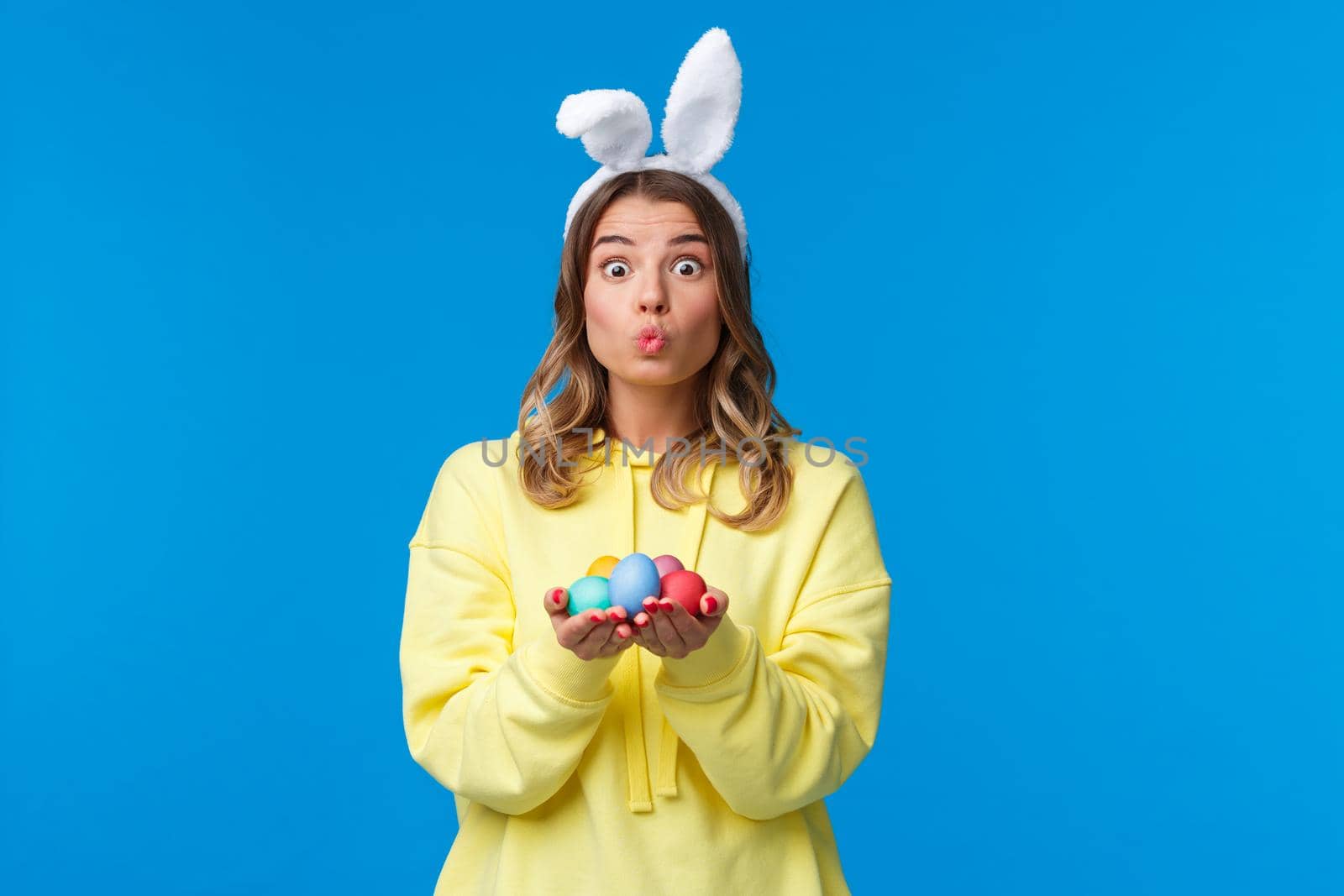 Holidays, traditions and celebration concept. Silly cute caucasian blond girl present you Easter eggs, painted it for holiday, show mwah kiss expression and wear rabbit ears, blue background.