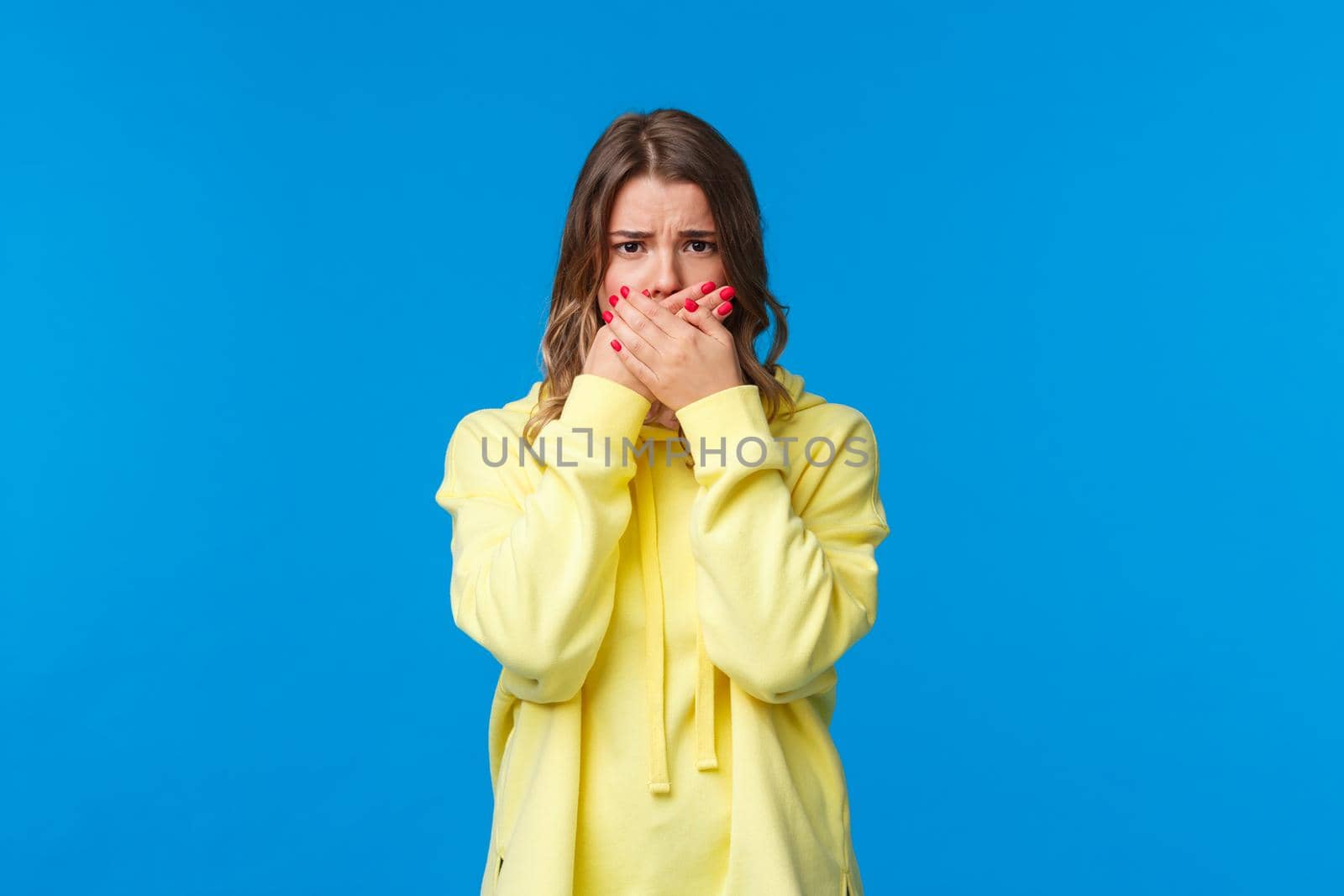 Upset and uneasy cute gloomy teenage girl have pimple near lip, cover mouth and frowning complaining, being offended unwilling to speak person who insulted her, stand blue background.