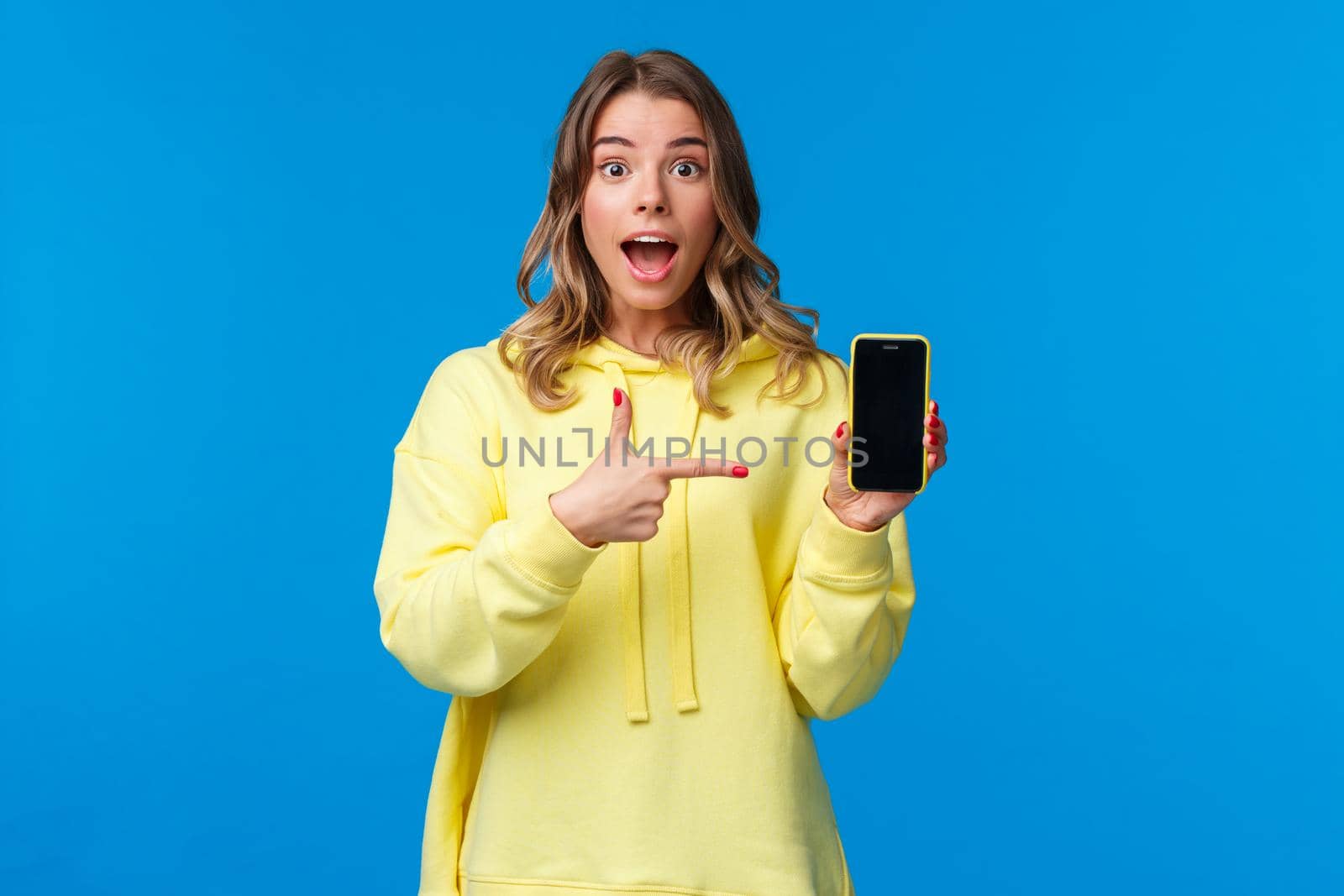 Impressed happy cheerful blond girl explain something as pointing mobile phone display, talking about new application or sharing her vacation photos, hold smartphone, look camera.