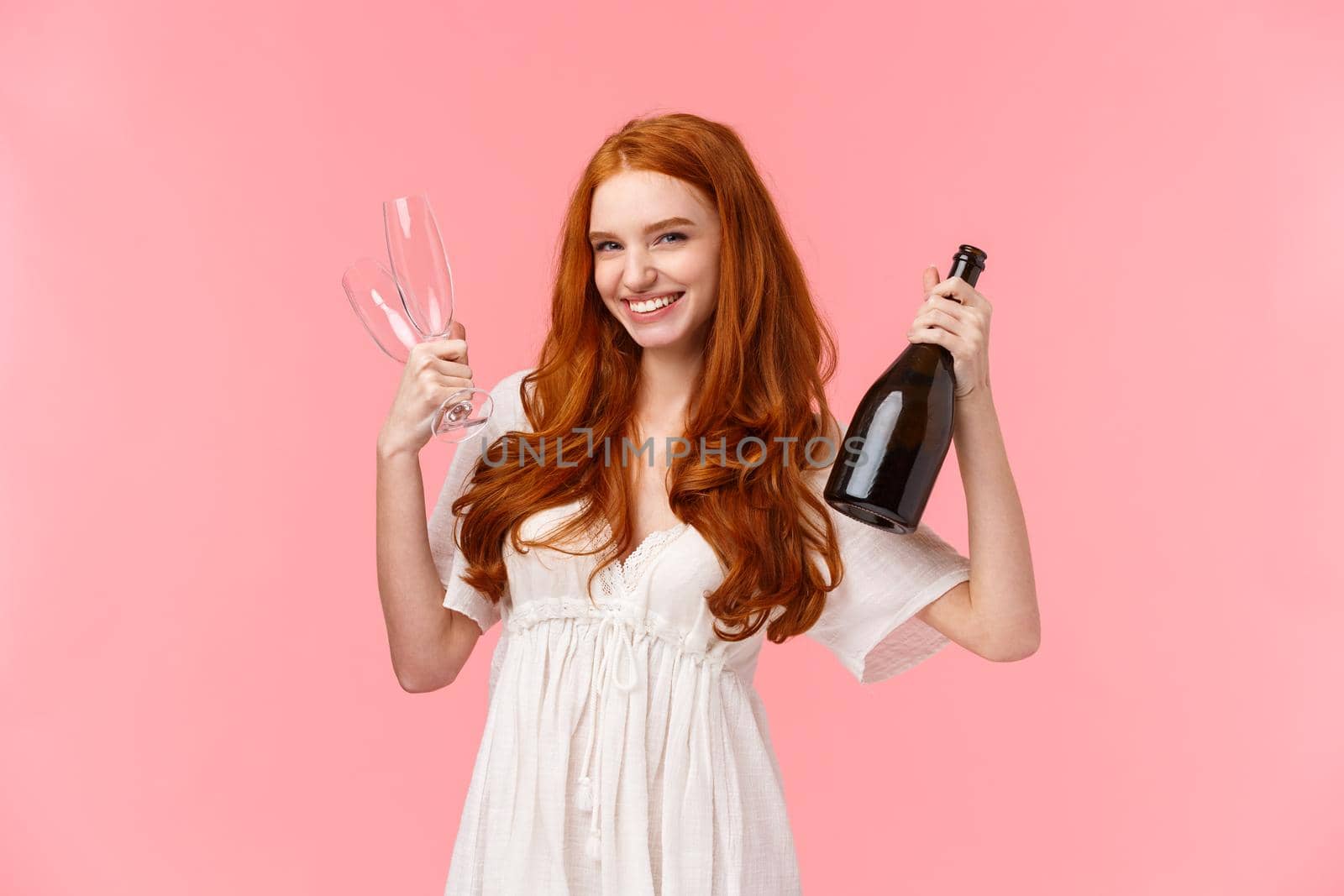 Sassy good-looking redhead woman celebrating with girlfriends, having fun together, holding bottle champagne and two glasses, invite for drink, smiling cheeky and happy, stand pink background.