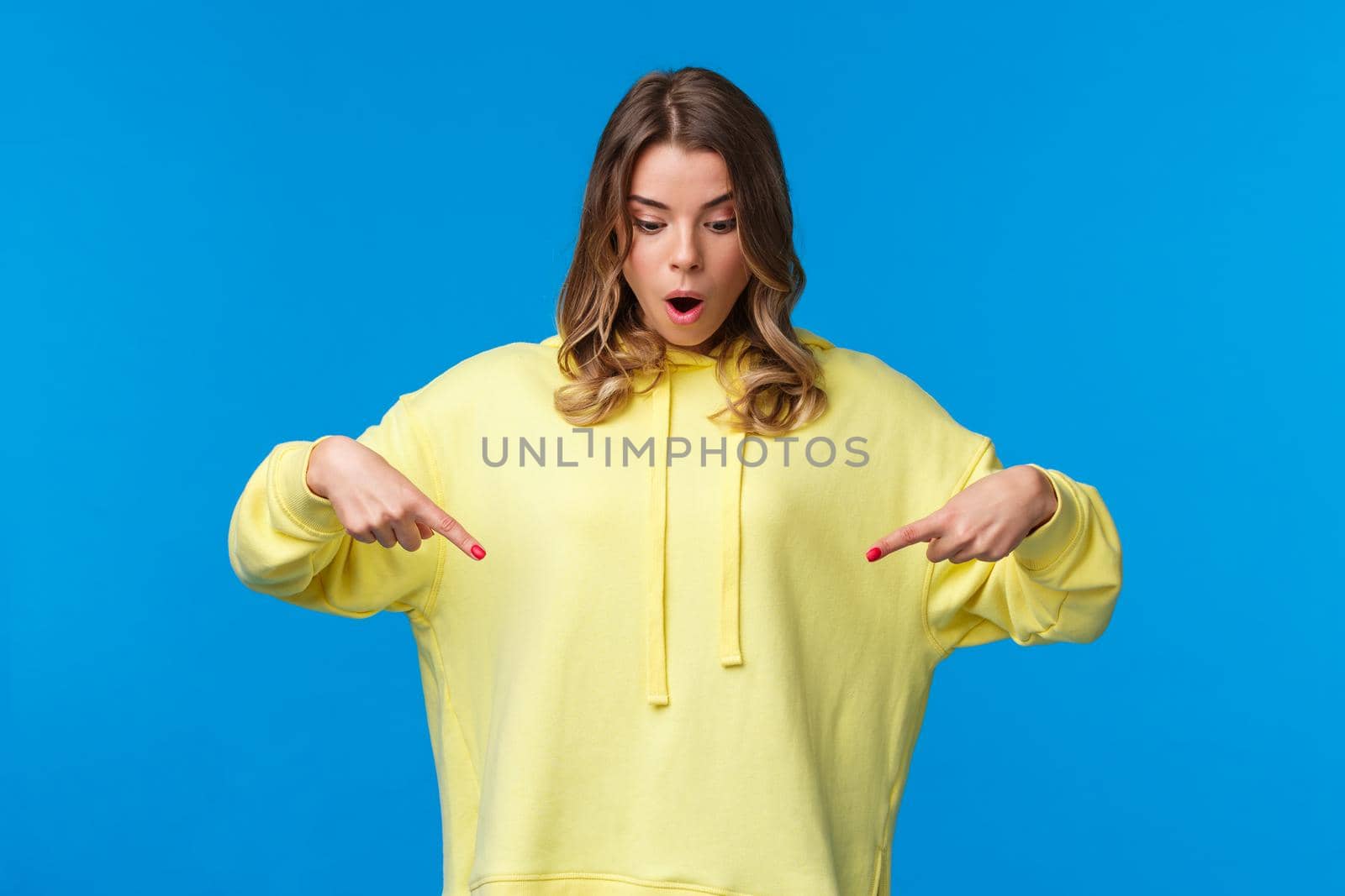 Girl found something stunning. Amused and speechless surprised blond alluring woman in yellow hoodie, drop jaw stare and pointing fingers down at cool product, blue background.