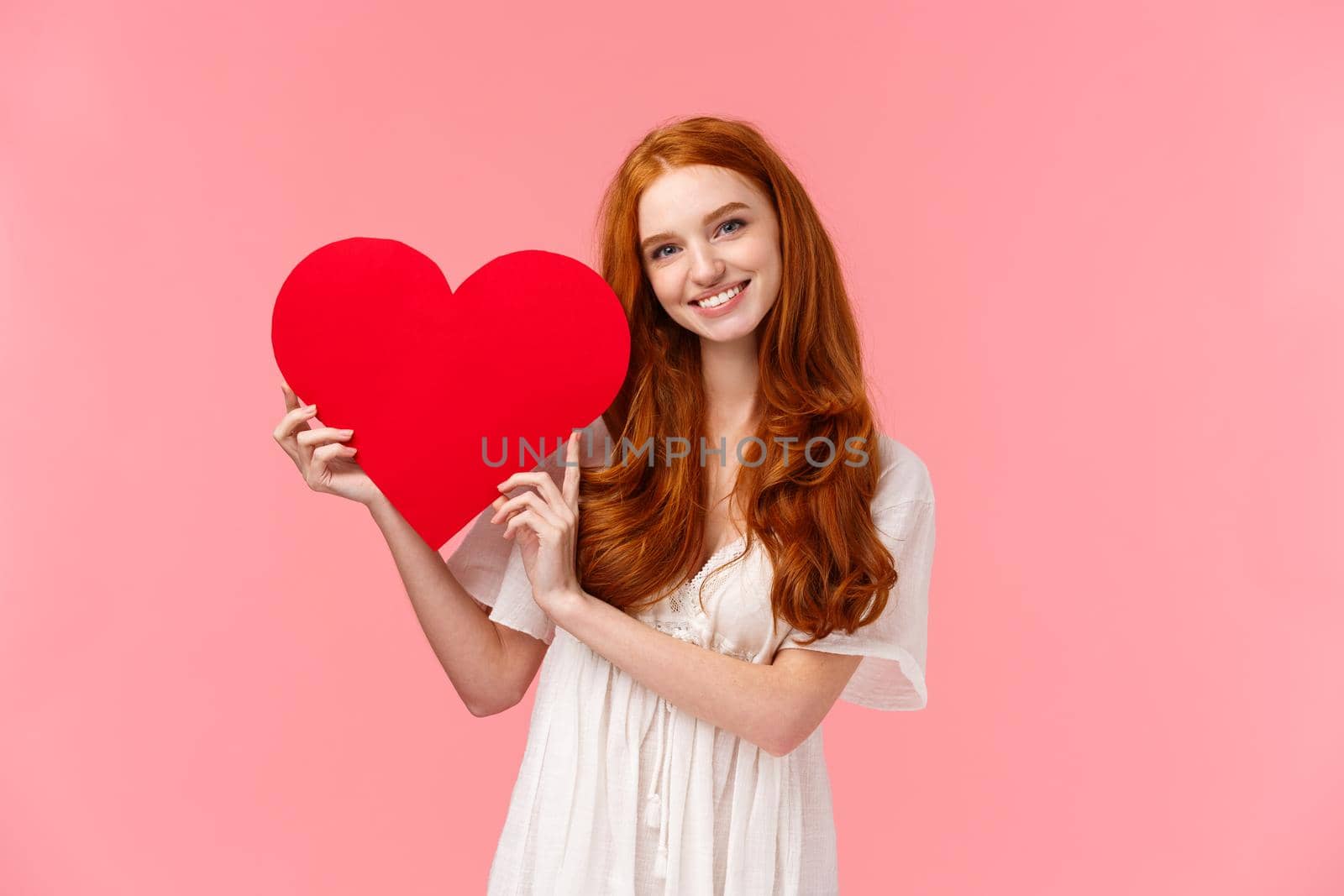 Relationship, care and valentines concept. Tender lovely redhead caucasian female in white dress, holding big red heart and smiling, confessing love, express sympathy or admiration, pink background.