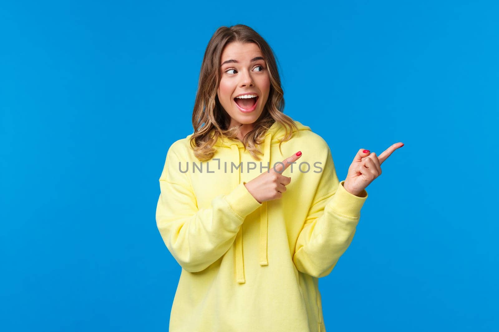 Excited happy, cheerful caucasian female with white beaming smile, wear yellow hoodie, react thrilled and amused to awesome news, cool banner, pointing fingers right, look away, blue background.