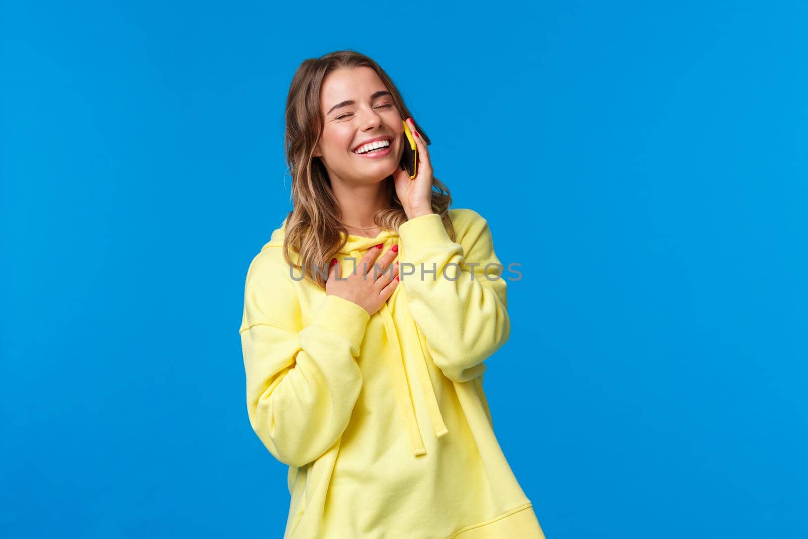 Waist-up portrait of joyful smiling young woman having funny conversation on phone, holding smartphone near ear, close eyes and laughing as touch chest, standing blue background.