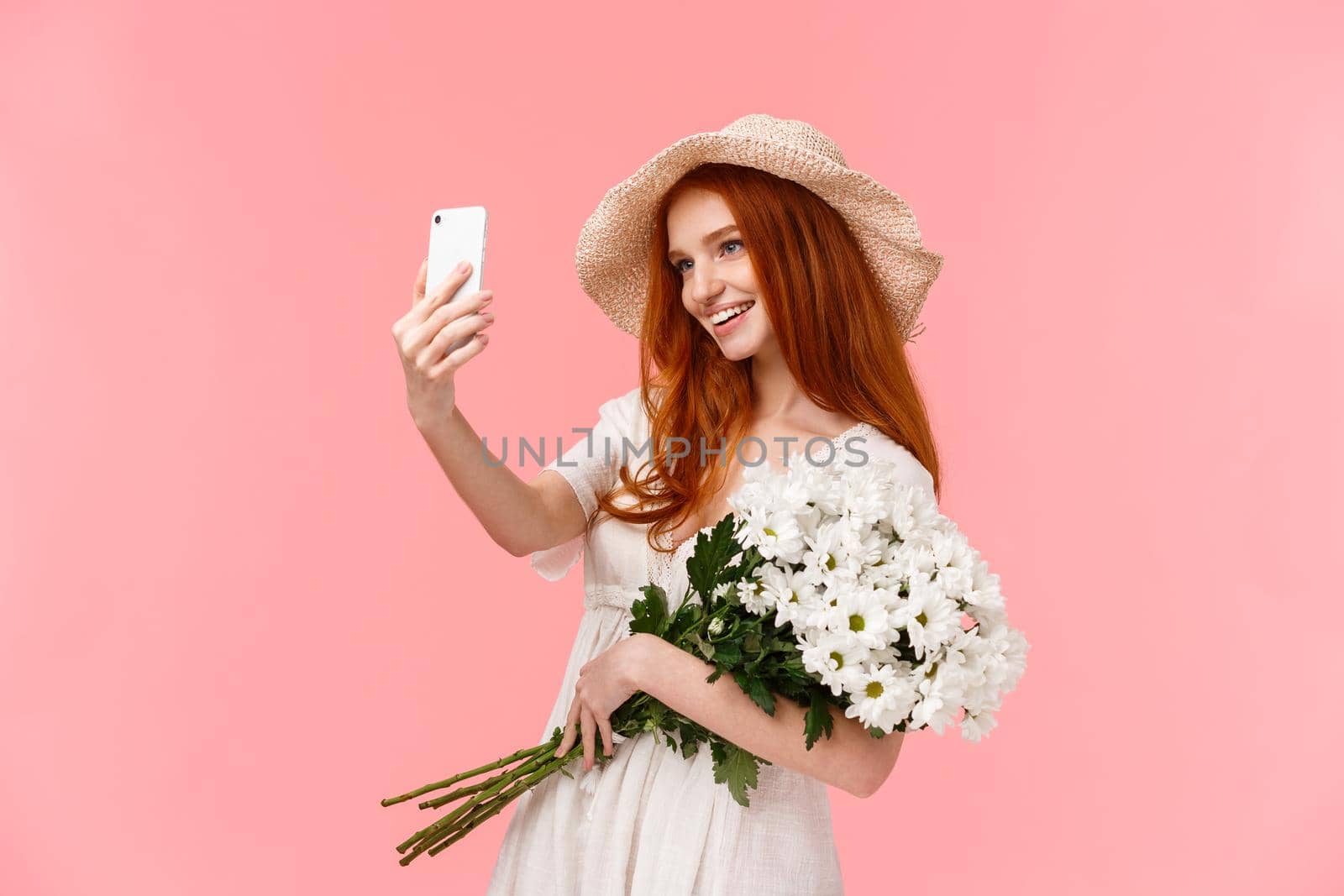 Celebration, social media and internet concept. Alluring sassy redhead female in straw hat, spring dress, holding bouquet, taking selfie on smartphone with white flowers, smiling pleased.