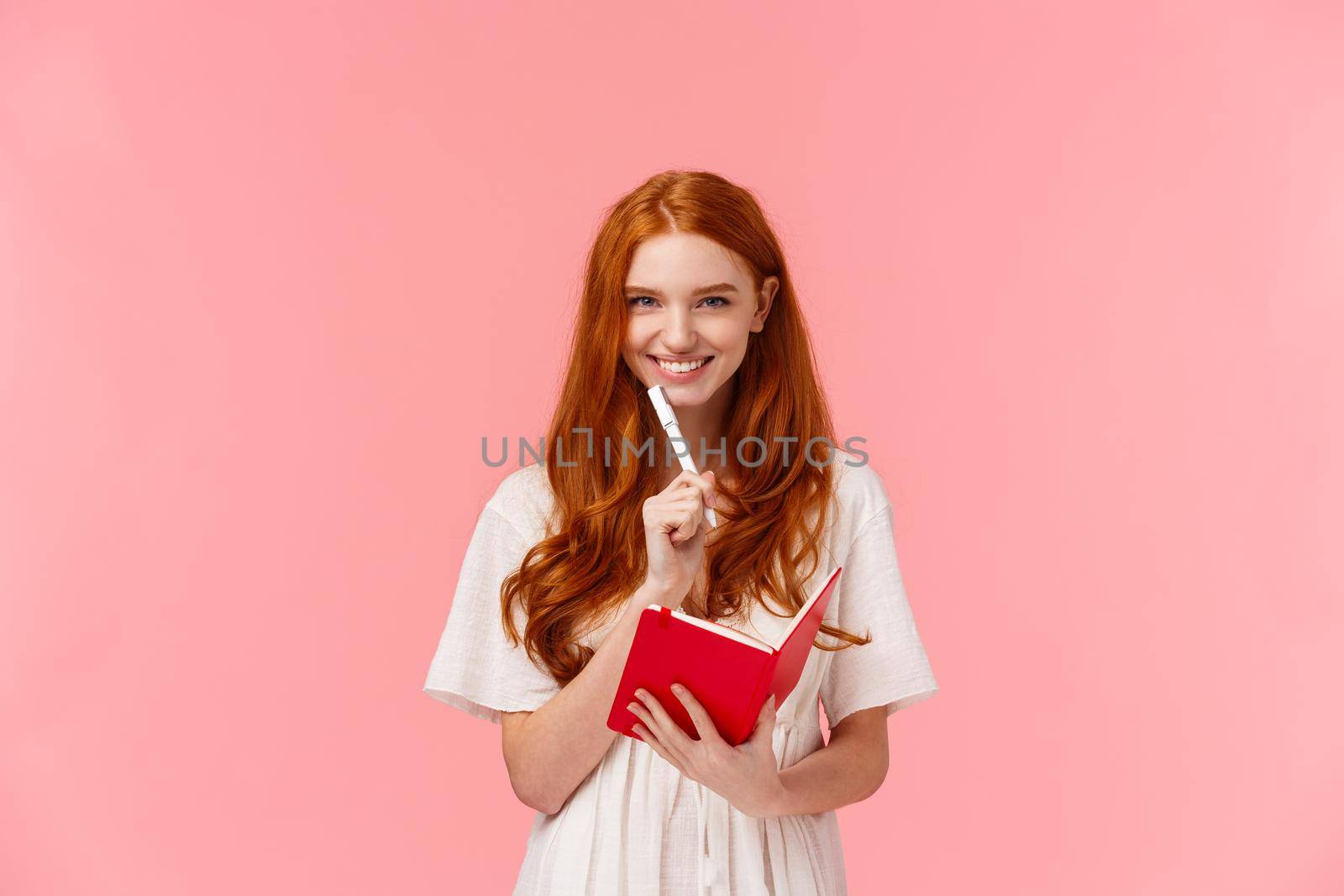 Excited cheerful redhead girl working, taking notes during lecture or attending courses, drawing something, smiling confident and pleased, touching face with pen, holding red notebook.