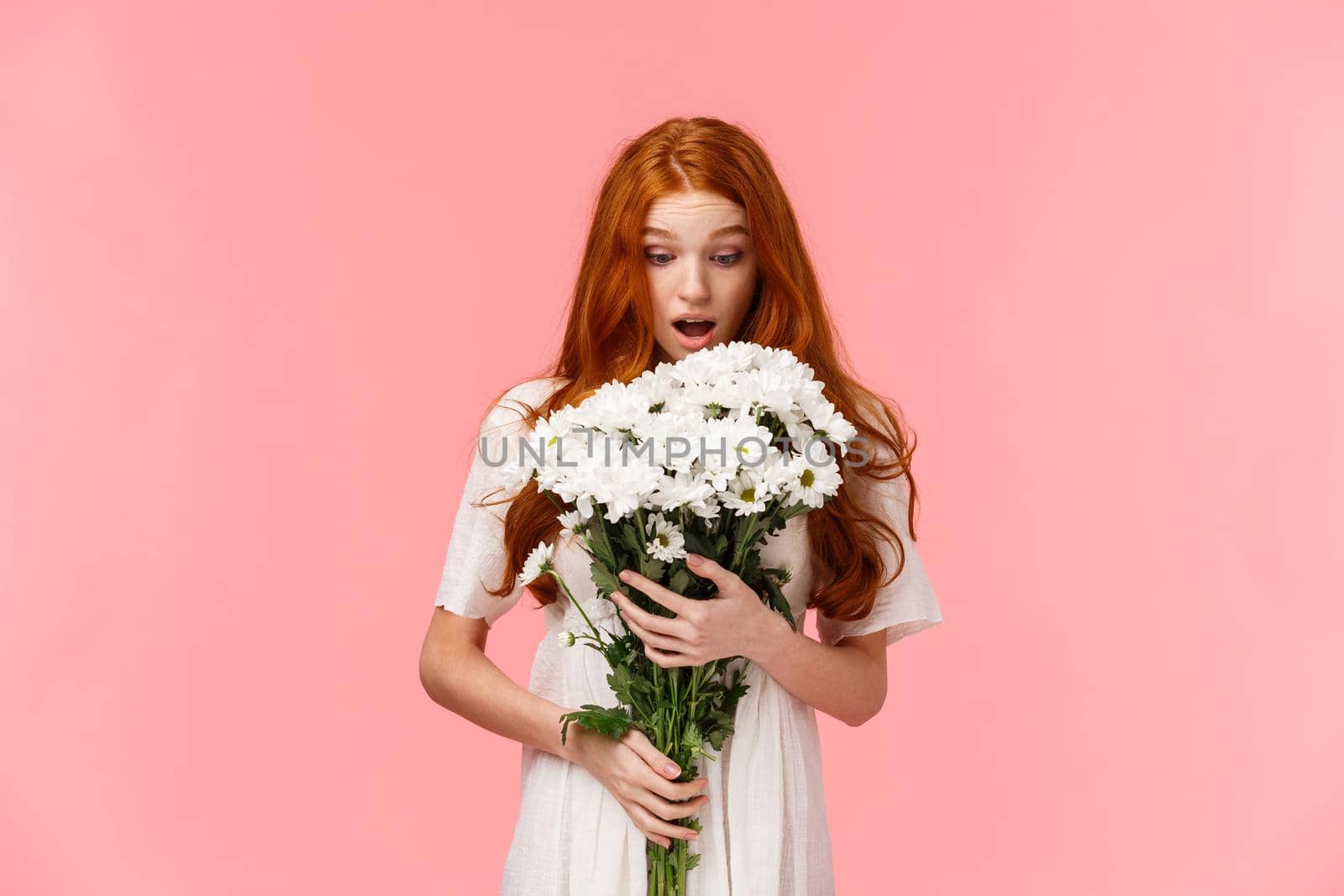 Surprised attractive redhead girl receive romantic valentines day gift, looking at beautiful bouquet of flowers amazed and wondered who might sender be, standing over pink background.