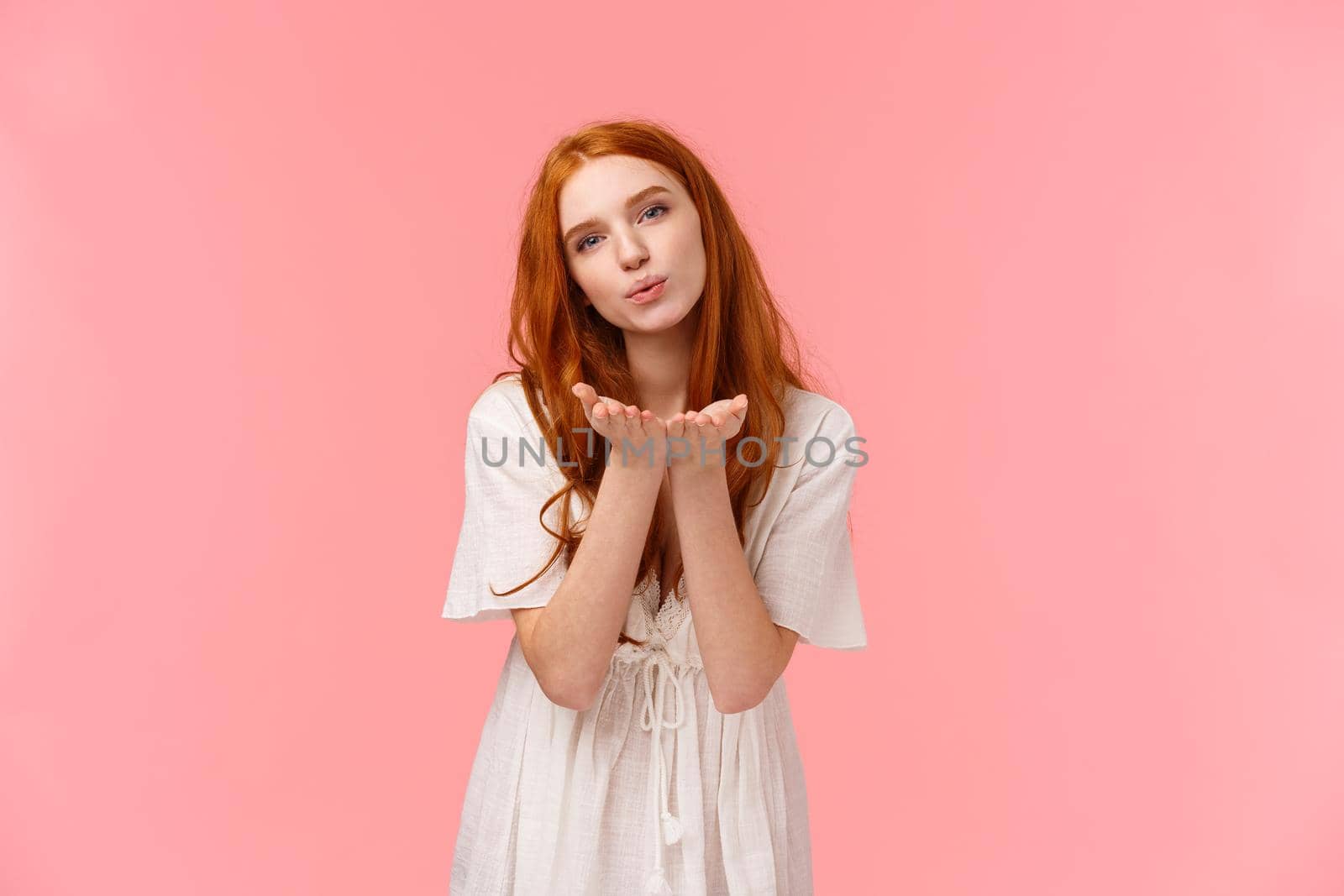 Waist-up portrait, cute and sensual, romantic feminine girl with red curly hair in white dress, bending and holding palms near folded lips, sending air kiss at camera, standing pink background.