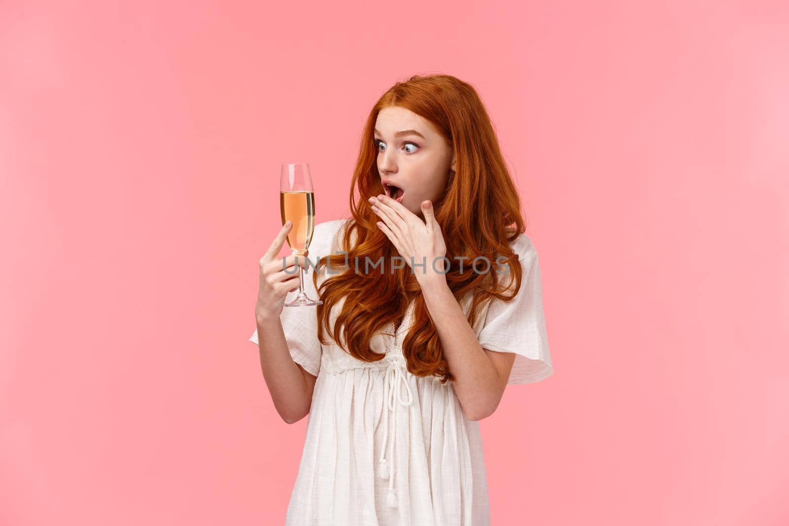 Shocked and astounded redhead woman surprised as cant remember when she took glass with champagne, have drinking problems, realise drank too much on party, standing pink background.