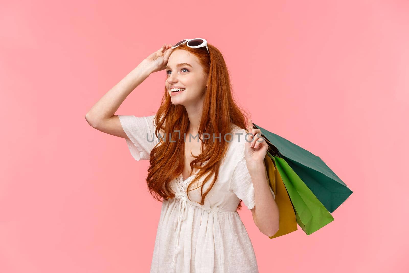 Amused and curious young redhead girl shopping stable upon interesting goods in store, looking intrigued and interested, smiling as wonder across shop mall, holding bags behind bag, take-off glasses.