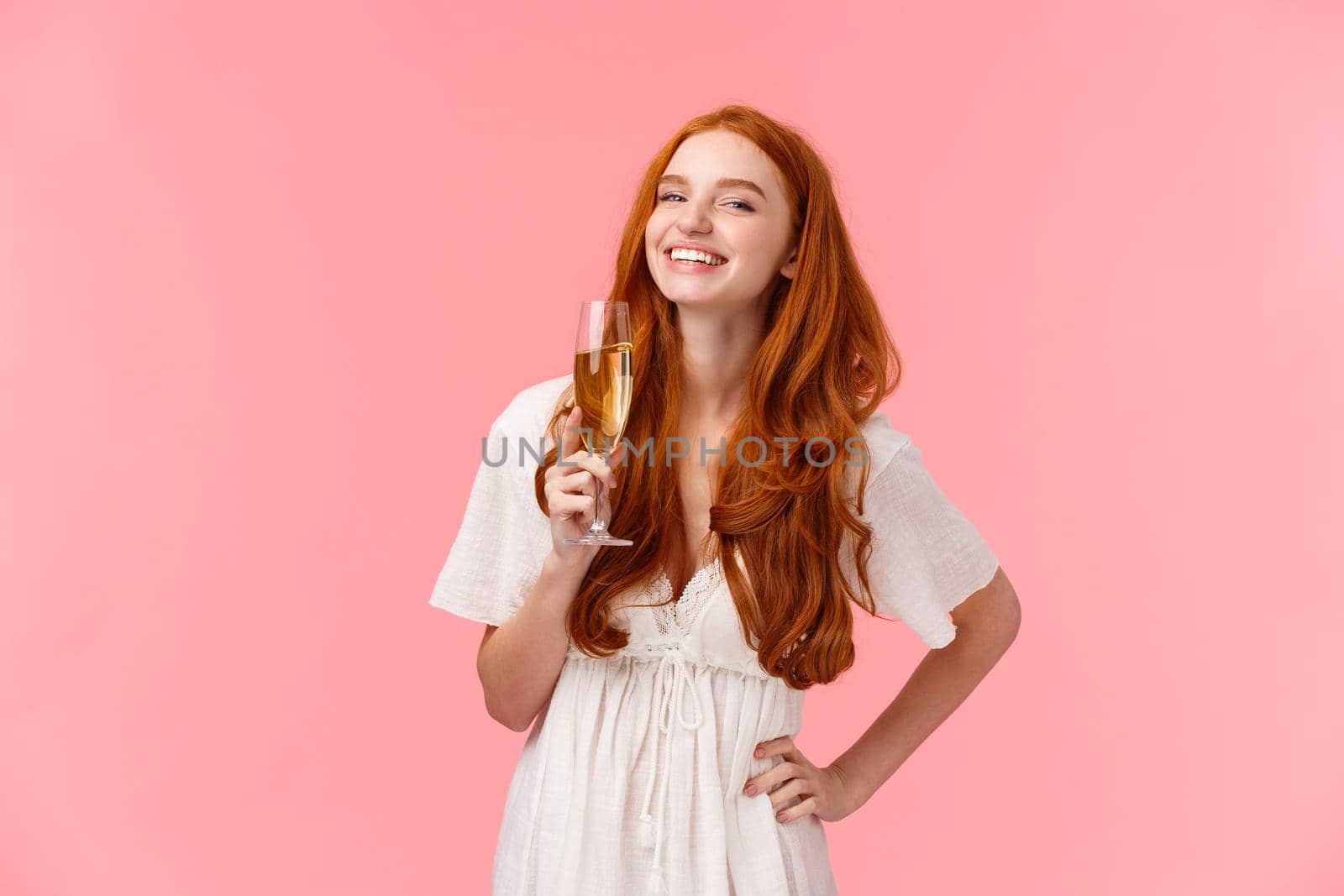 Happiness, confident and celebration concept. Happy and carefree alluring redhead b-day girl sipping champagne and laughing, having fun, enjoying party, standing over pink background.