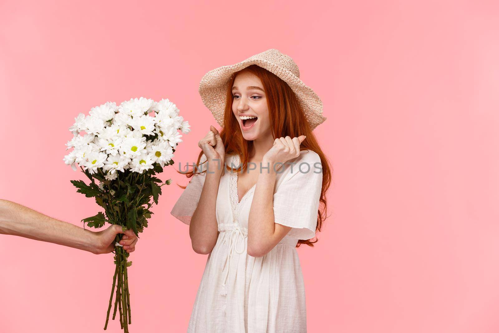 Happy moments, romance and relationship concept. Joyful and surprised cute, lovely redhead girl jumping from happiness and amazement, looking at hand giving her beautiful flowers, pink background.