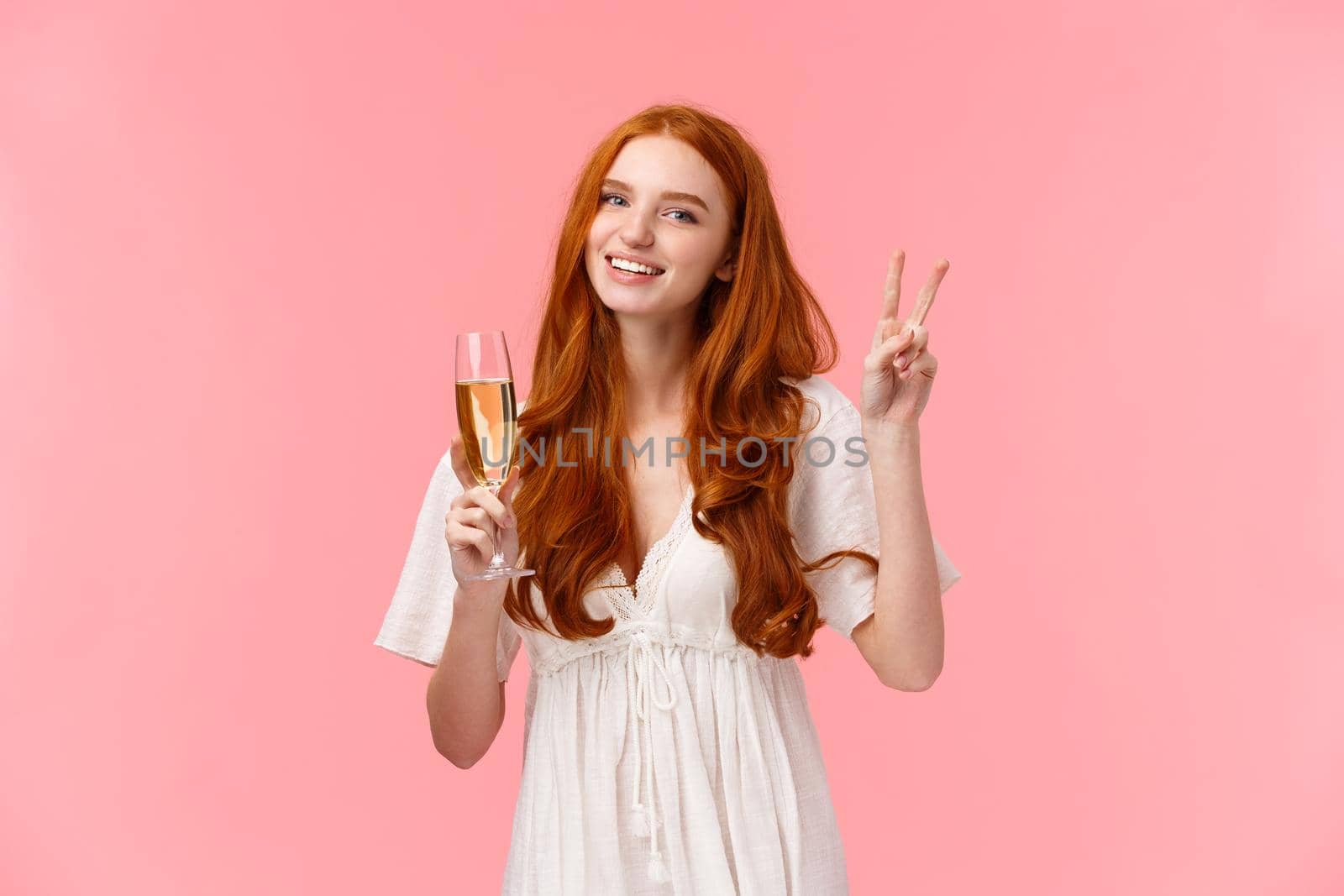 Lovely romantic caucasian redhead female in white cute dress, showing peace sign, informal greeting of friend on party, smiling holding glass of champagne, drinking as celebrating, attend event.