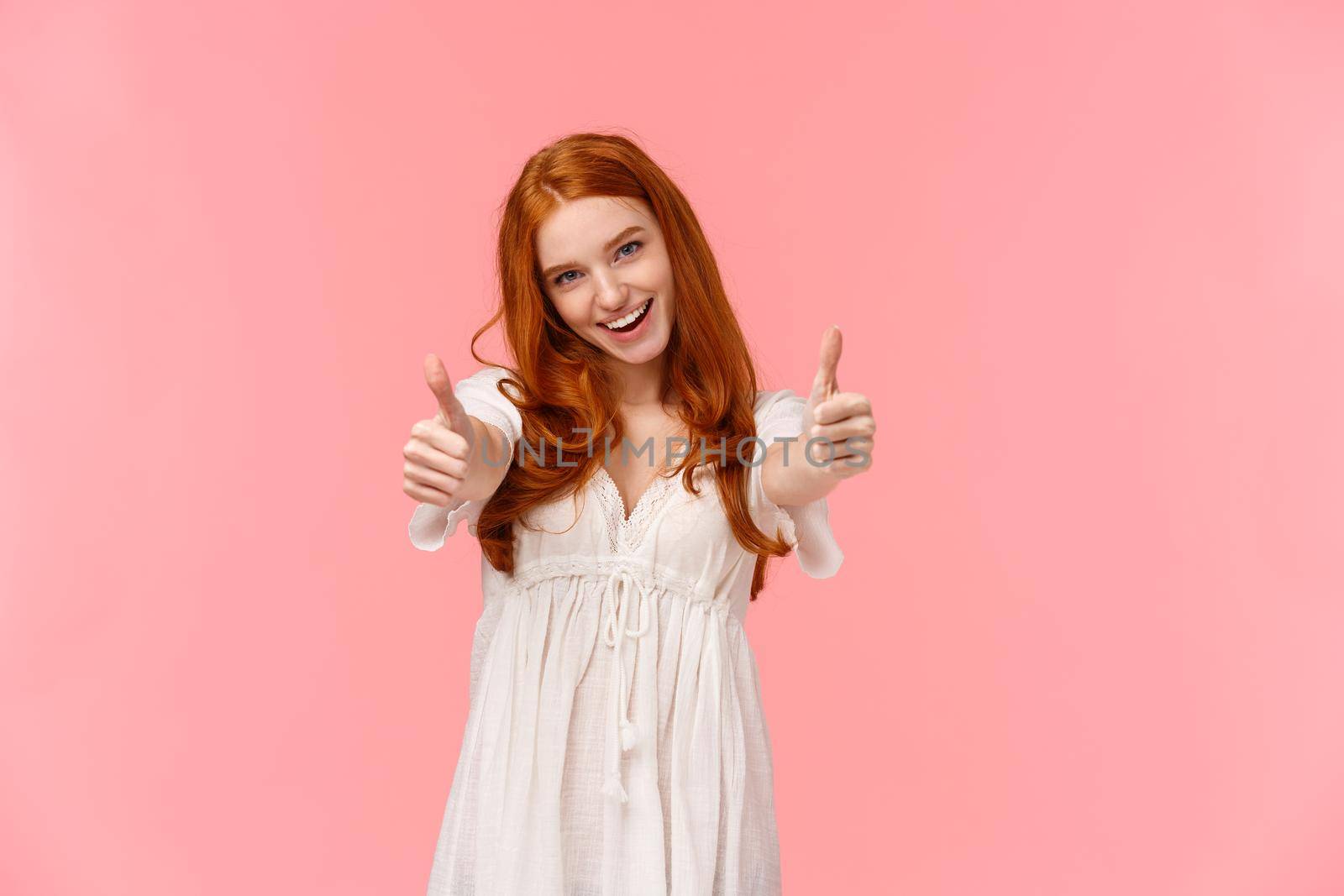 I support your choice. Charismatic excited, happy redhead woman in white dress, showing thumbs-up to cheer you, agree or express satisfaction of cool product, smile approval, pink background.