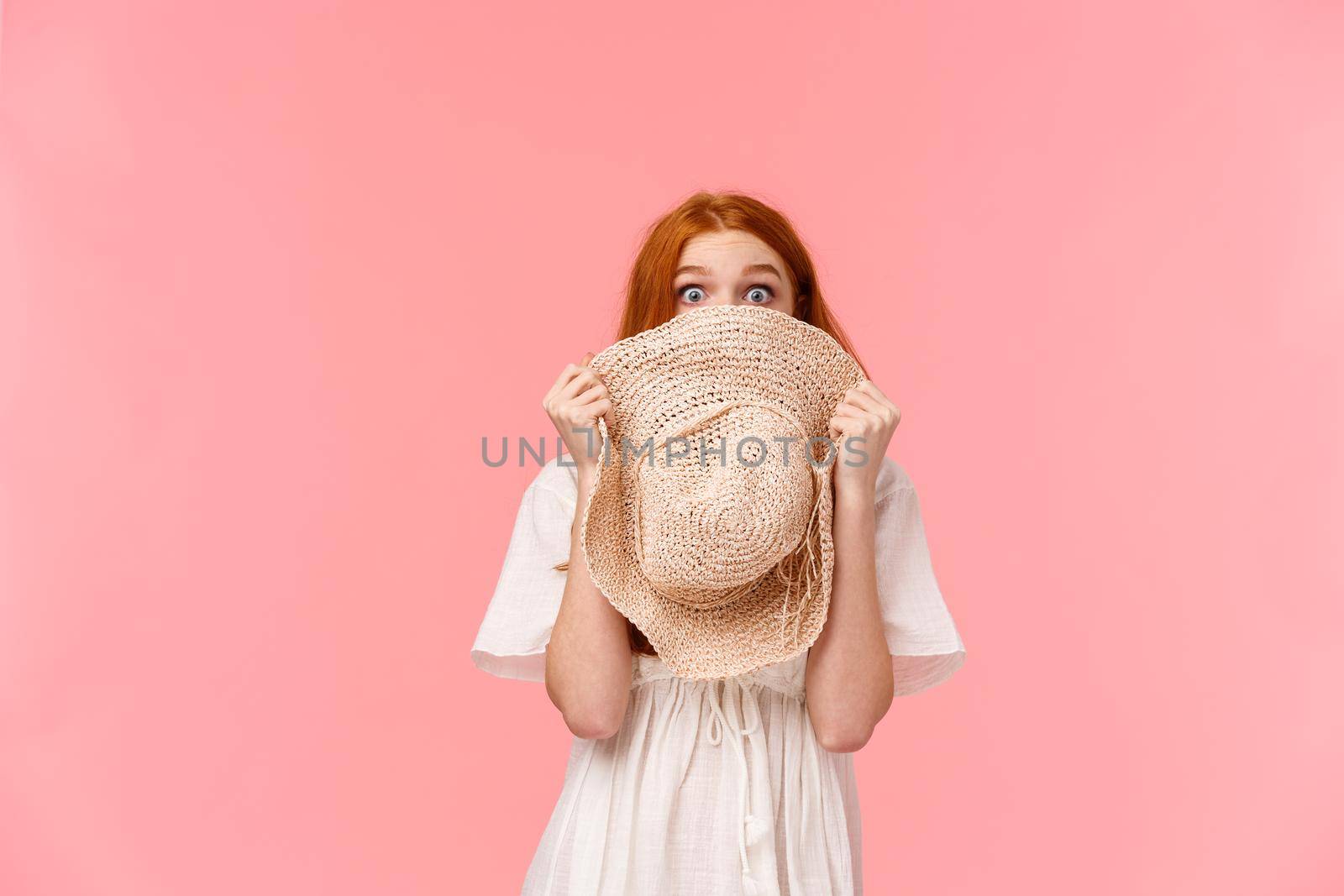 Surprise, look at camera. Shy and blushing cute startled redhead girl hiding face behind straw hat, staring camera scared or shook, standing pink background overwhelmed.