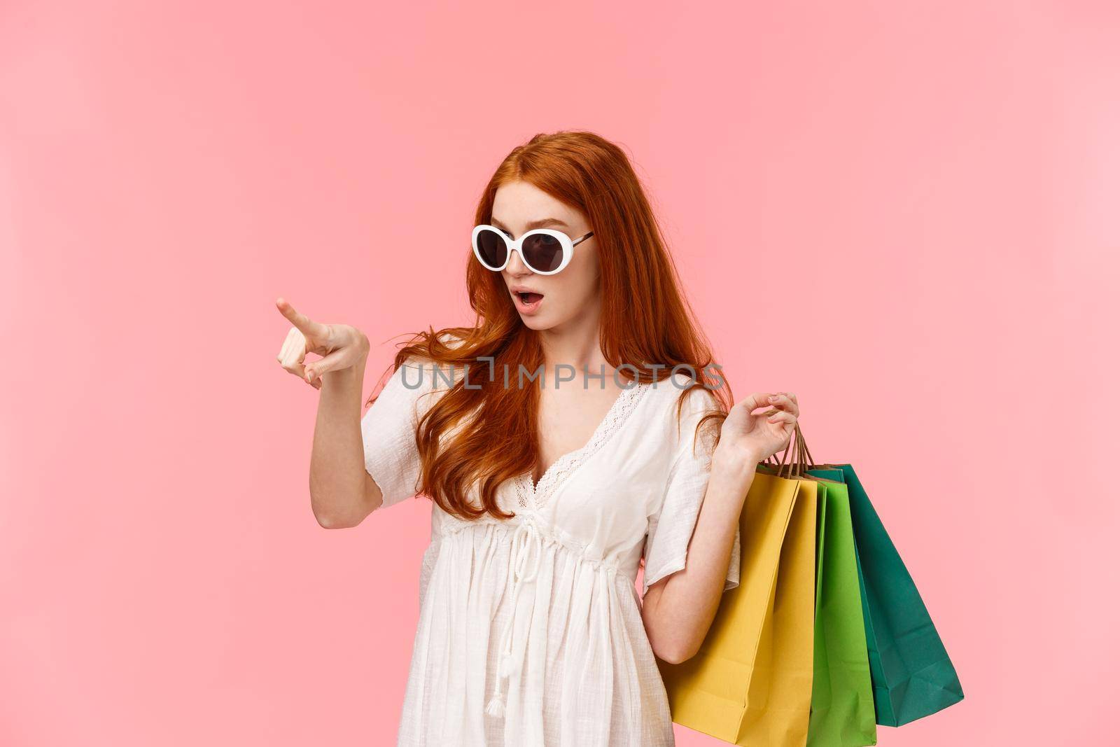 Girl showing girlfriends perfect dress for prom, checking out something interesting, pointing finger and heading to store to buy product, holding shopping bags behind back, standing pink background.