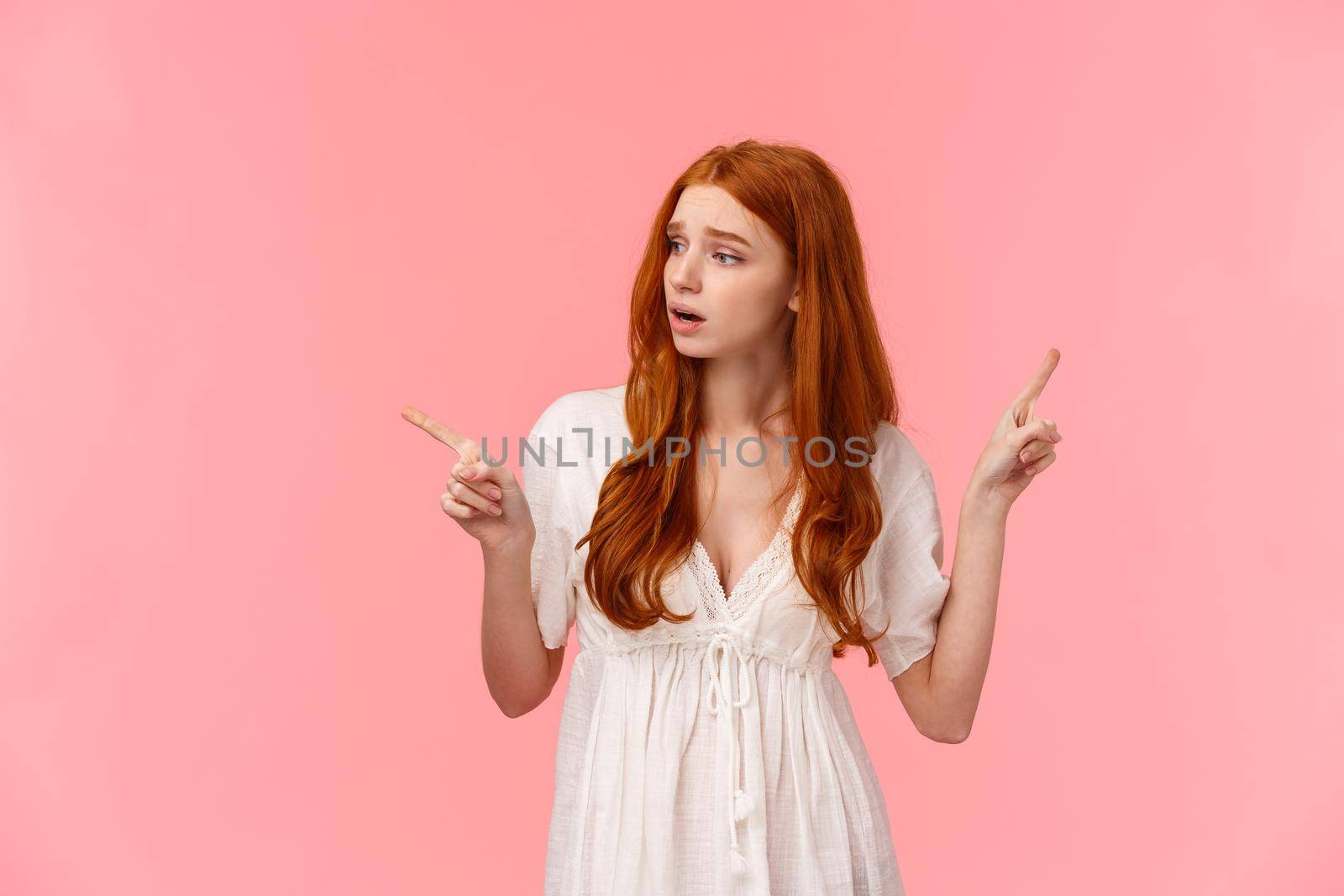 Insecure, worried cute redhead girl scared making wrong choice, look around indecisive and unsure, pointing sideways, indicate left and right product, frowning upset, pink background.
