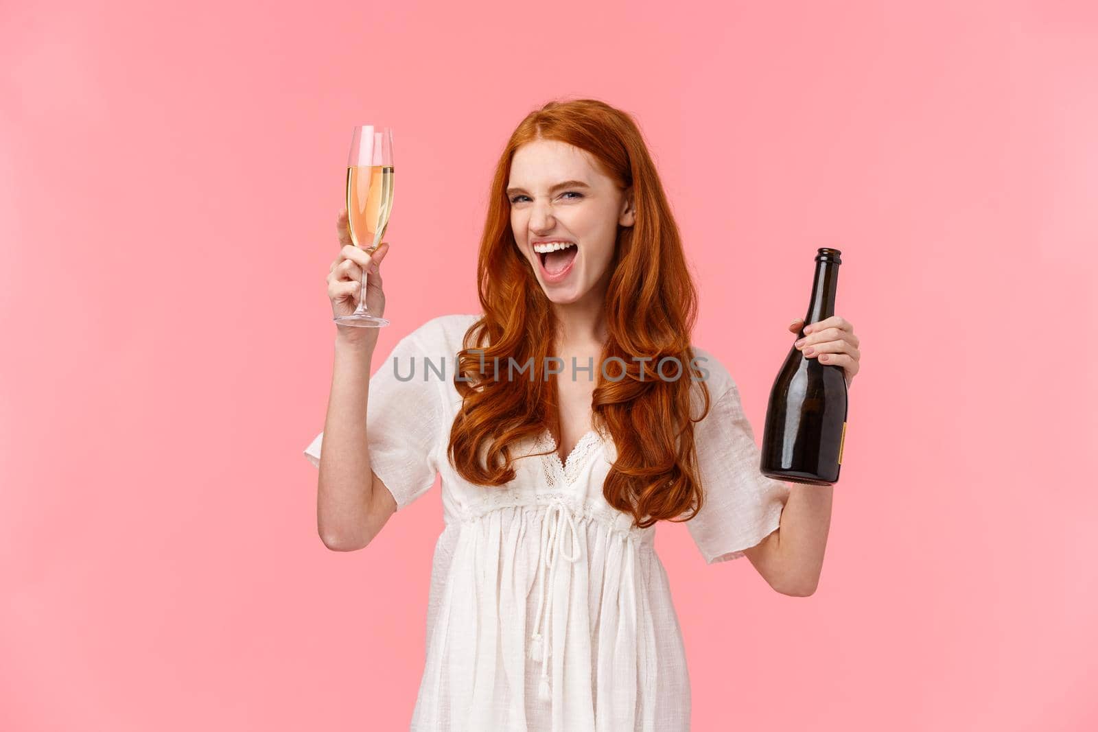 Excited and carefree drunk redhead caucasian girlfriend having fun, going wild and free on awesome party, raising glass and bottle champagne, smile sassy, stand pink background.