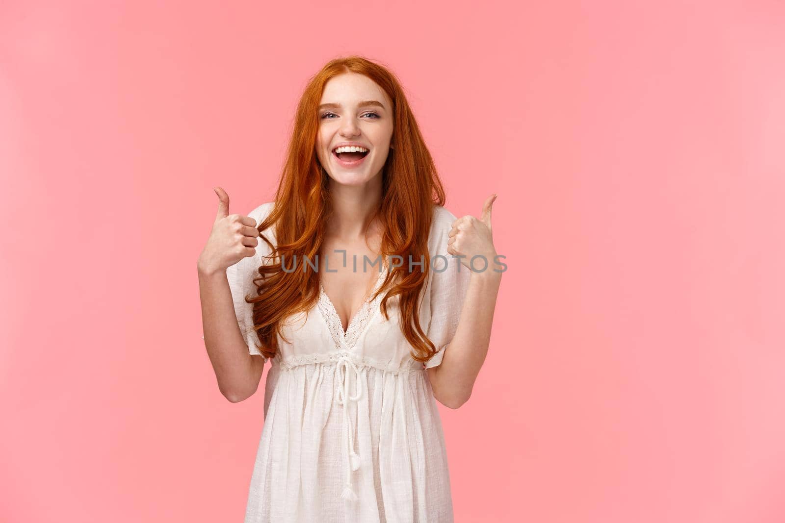 Waist-up portrait cheerful, upbeat redhead woman in white dress, showing thumbs-up and smiling in approval, like product, recommend it, express positive opinion, standing pink background.