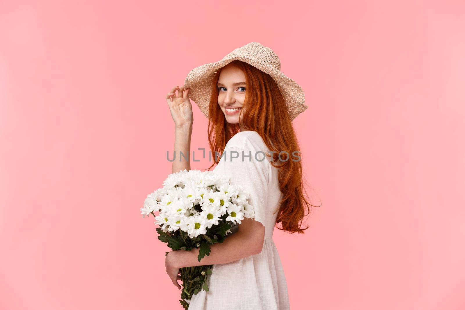 Romantic, silly and tender feminine redhead woman in cute hat, dress, holding bouquet white flowers, turn camera and smiling coquettish, flirting with boyfriend over pink background.