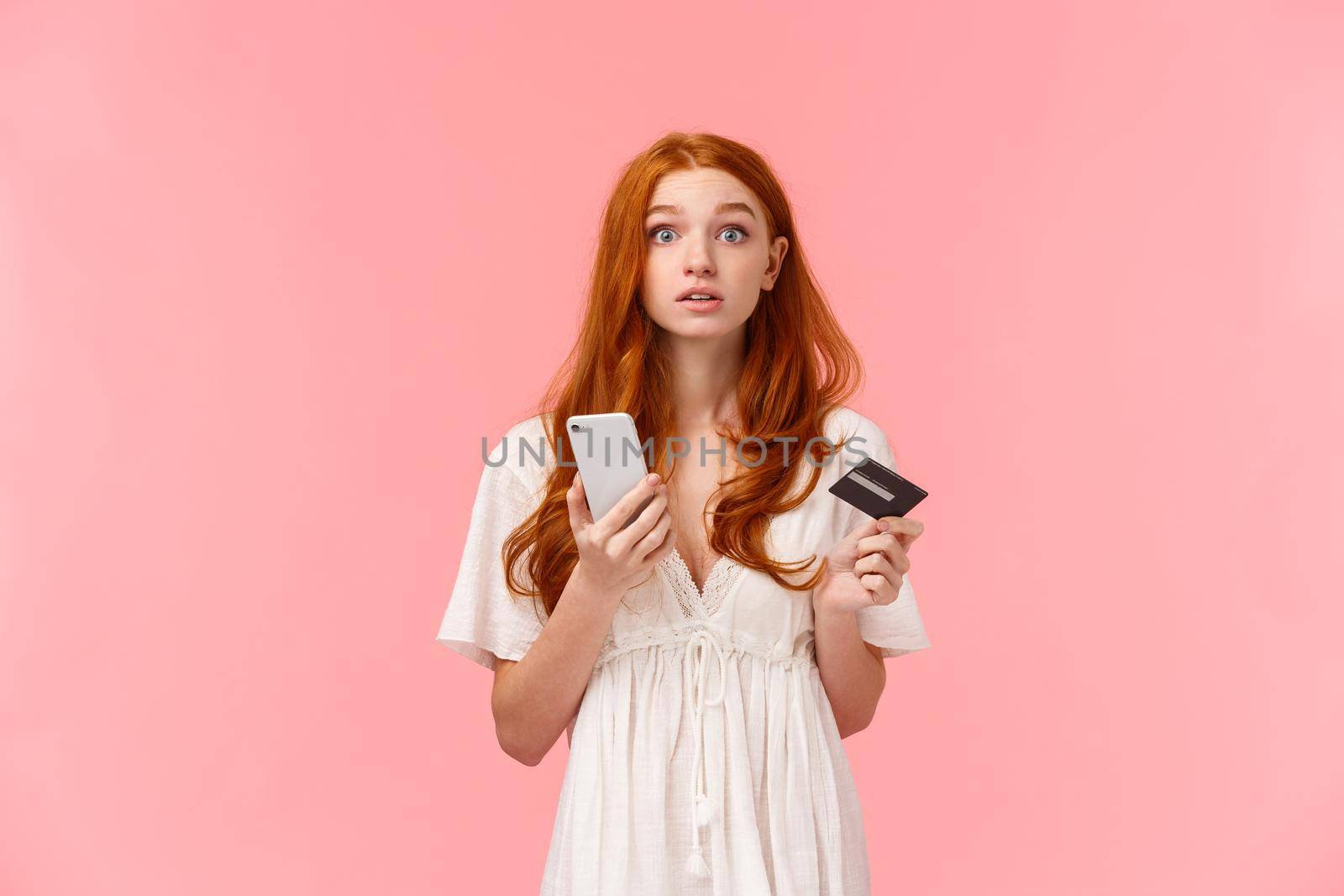 Distracted questione cute redhead girl raise head at camera to answer question, looking wondered camera, holding smartphone and credit card, trying pay for online purchase cant understand something.