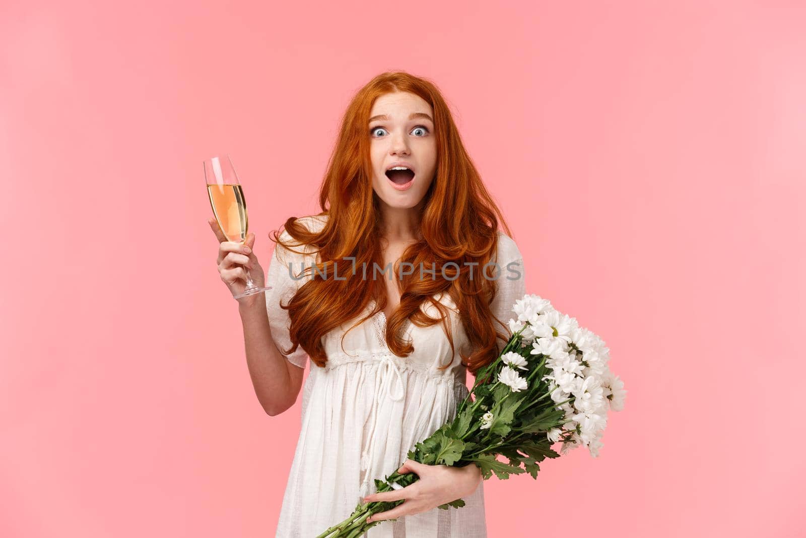 Excited and touched amused birthday girl receiving congratulations, look fascinated and surprised at camera, open mouth wondered, holding glass champagne and bouquet flowers, pink background.