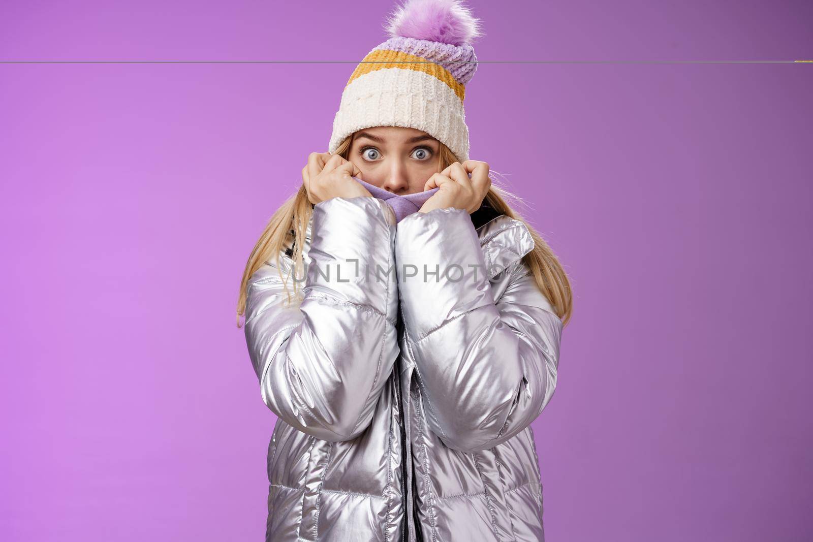 Afraid shocked young charming sister pulling jacket face cover hiding frightened scared terrifying stories snowman walking mountains widen eyes concerned look camera fear, standing purple background.