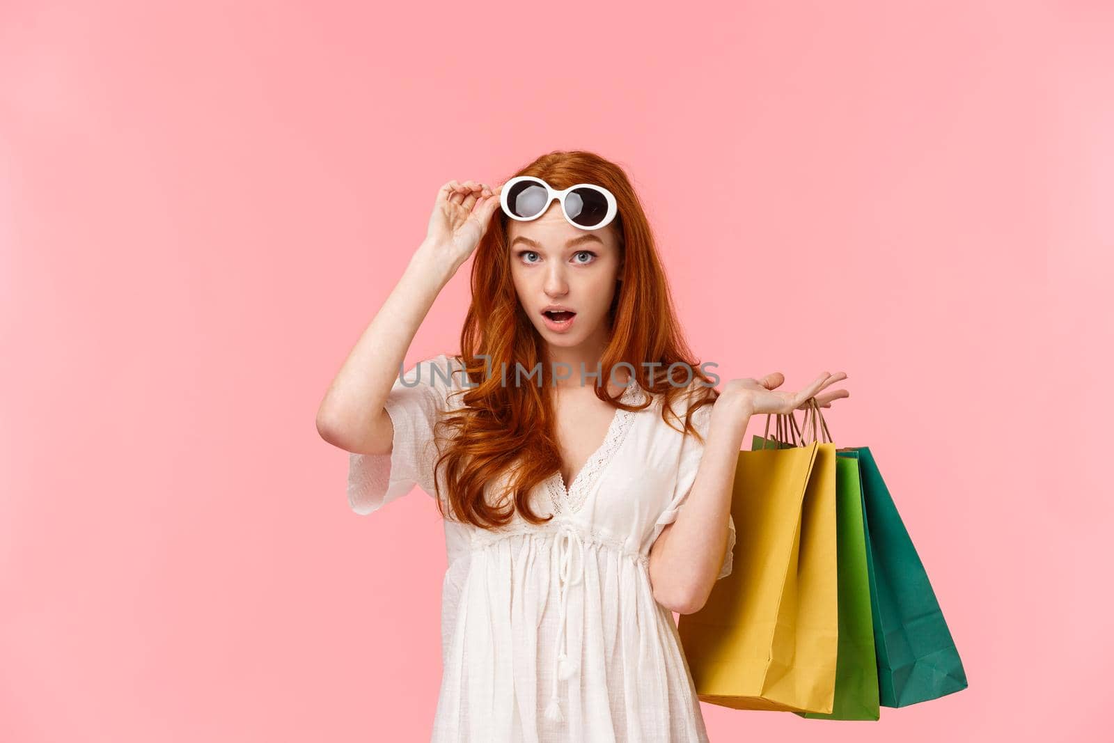 What an audacity. Shocked and speechless, astonished redhead girl take-off glasses to see something impressed, drop jaw, gasping, carry shopping bags behind back, standing pink background.