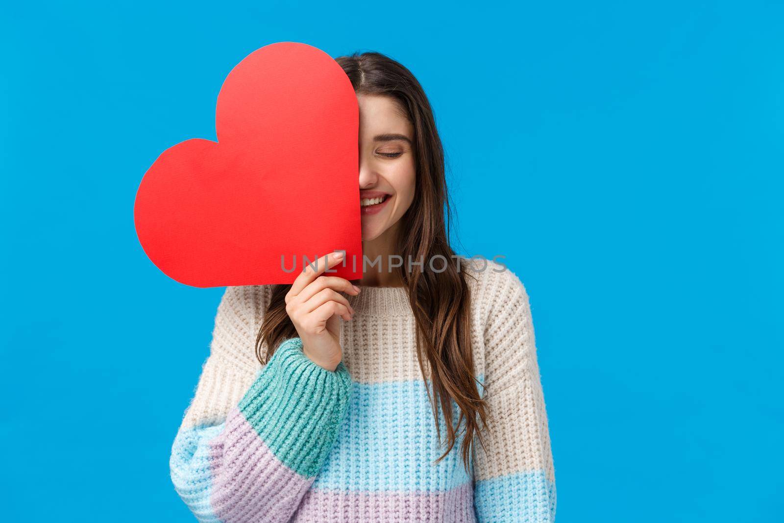 Happiness, relationship and love concept. Carefree happy good-looking woman holding big cute red heart sign over half face, laughing with closed eyes, blushing and feeling loved, blue background.