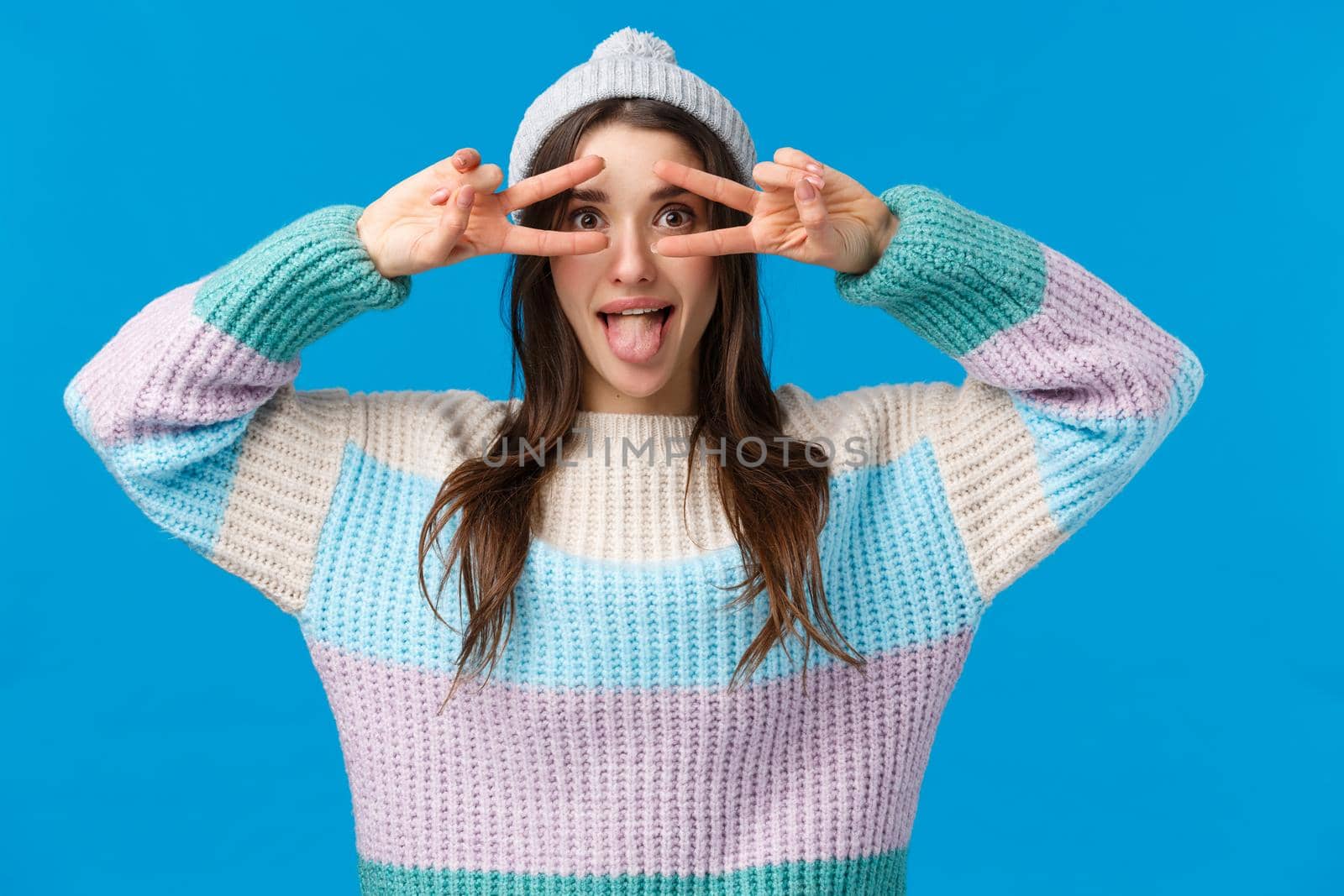 Be yourself. Attractive and playful funny, beautiful caucasian girl in winter hat, sweater, showing tongue and make disco, peace signs over eyes, having fun on christmas holidays, blue background.